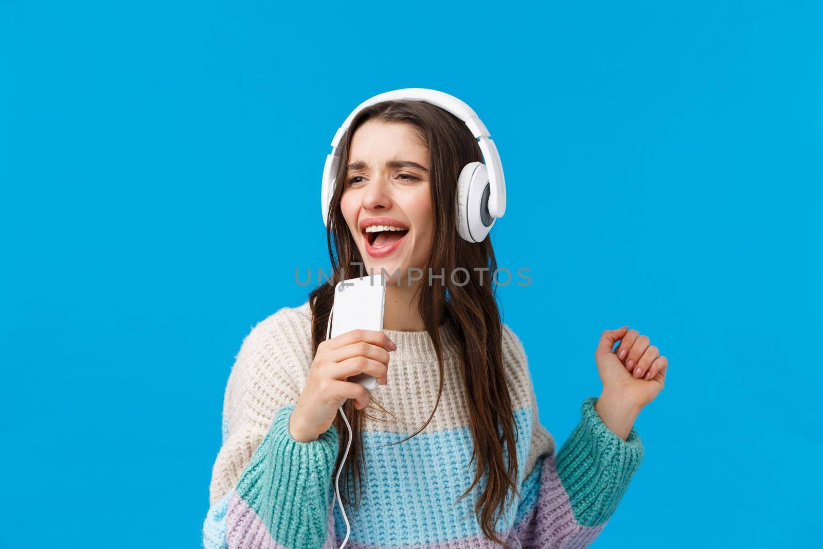 Waist-up portrait carefree good-looking young brunette woman playing karaoke game on phone, holding smartphone like mic, singing into phone dancing, listen music in headphones, blue background.