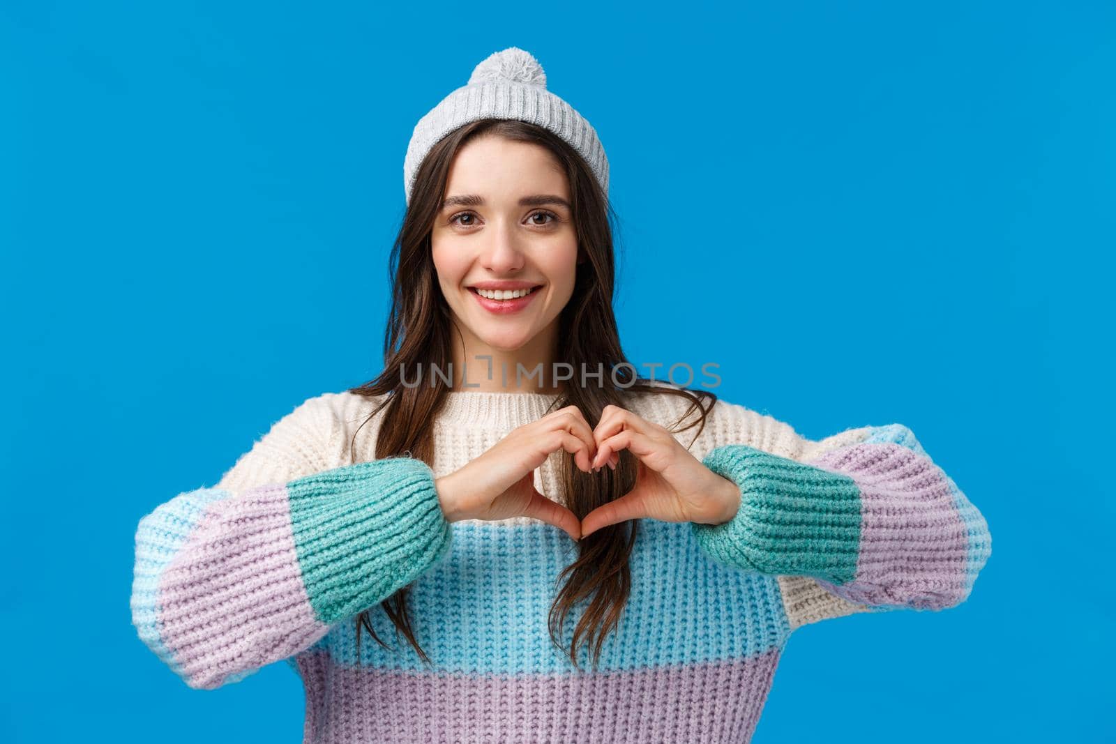 Valentines day, relationship and emotions concept. Lovely, pretty brunette girl in winter hat, sweater, showing heart sign and smiling, looking camera happy, show love and affection, blue background by Benzoix