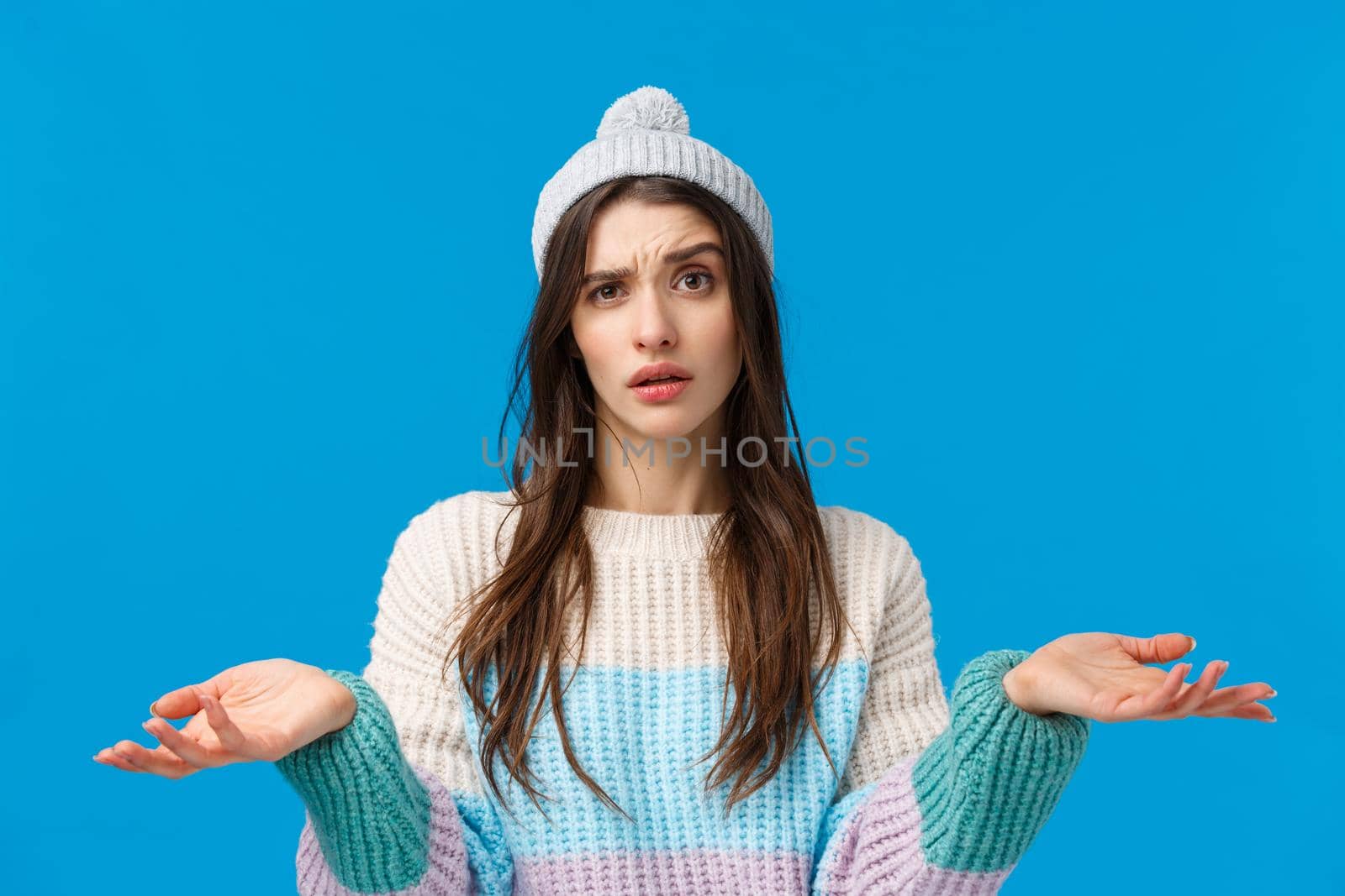 Waist-up portrait confused young attractive woman in winter sweater, hat, cant understand, standing clueless and uncertain, shrugging raise hands up dismay, dont know answer, blue background.