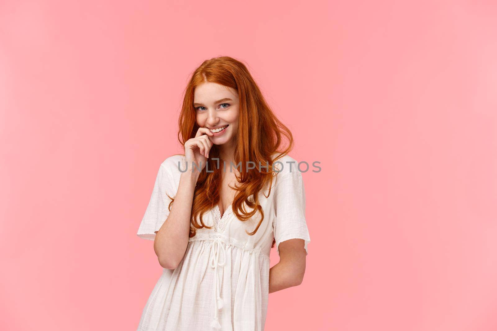 Romance, relationship and tenderness concept. Beautiful tender redhead woman in cute white dress, blushing and giggle silly, smiling biting finger gazing at camera sensual, flirt over pink background.