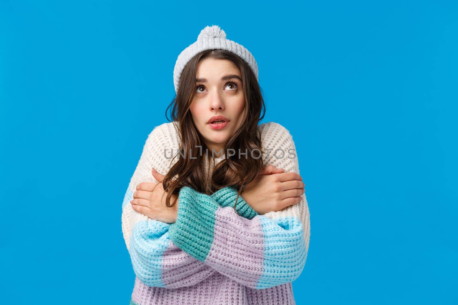 Girl watching at snowflakes, looking up and trembling from cold standing in winter hat, sweater, hugging herself to warm-up, embracing body clench teeth freezing temprature, blue background.
