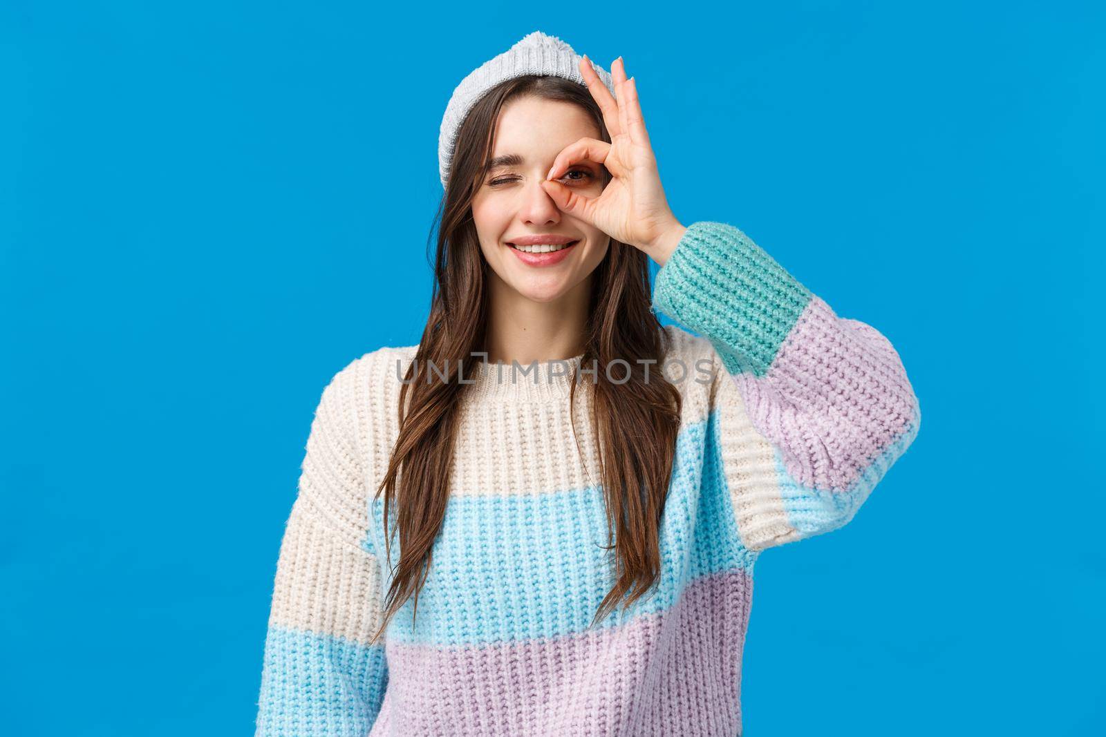 Waist-up portrait cheerful beautiful brunette woman in winter sweater, hat, showing okay, good gesture, OK sign over eye, smiling and wink in approval, assertive everything alright, blue background.