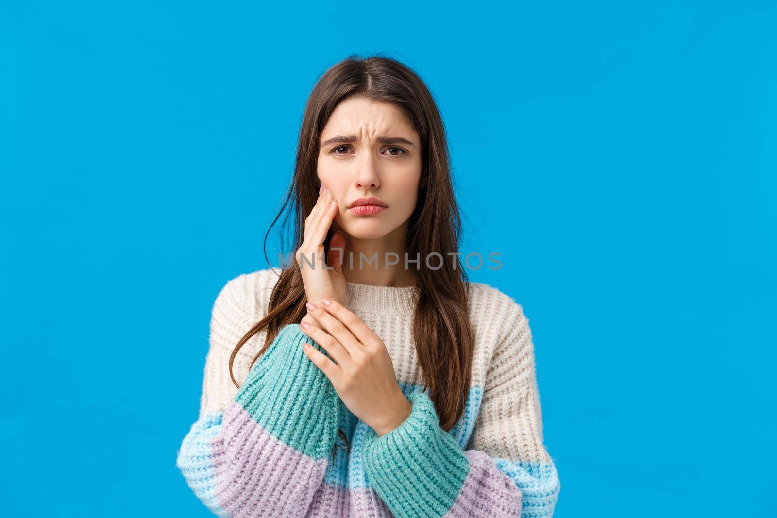 Waist-up portait sad silly girl having troubles with teeth, frowning squinting from pain, touching cheek as feeling toothache, have appointment dentist, standing blue background.