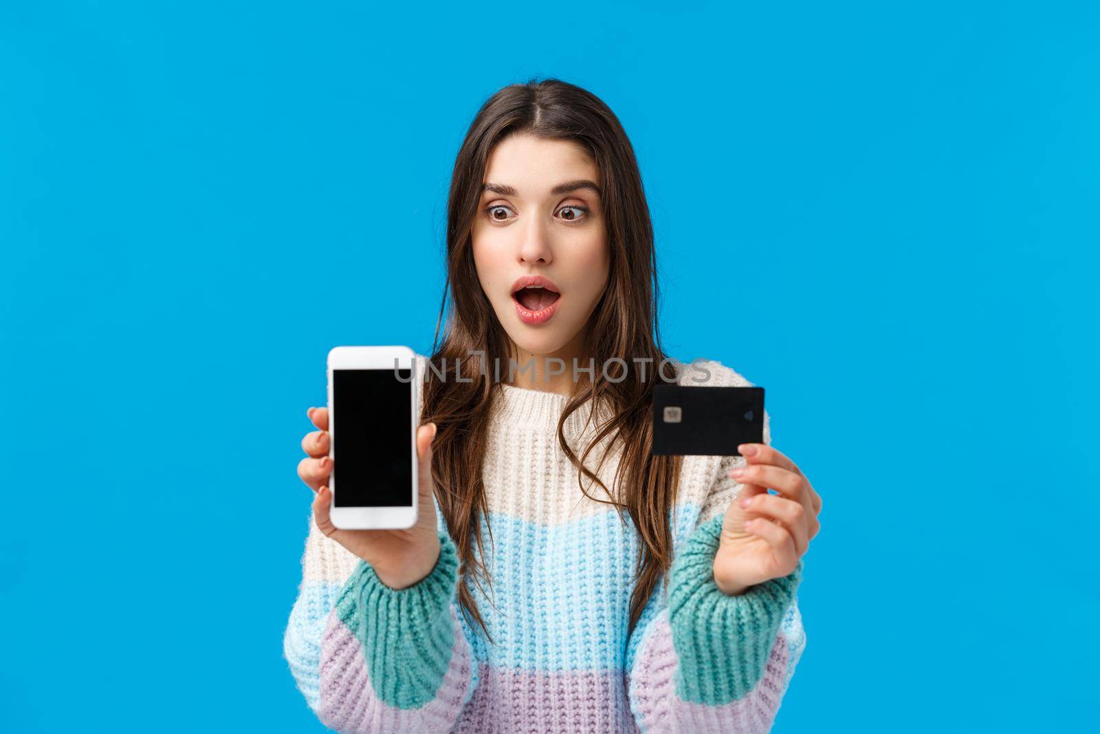 Excited, amazed speechless european woman react to something amazing, winter special offer from bank, shopping online, showing credit card and looking at smartphone, mobile display, blue background.