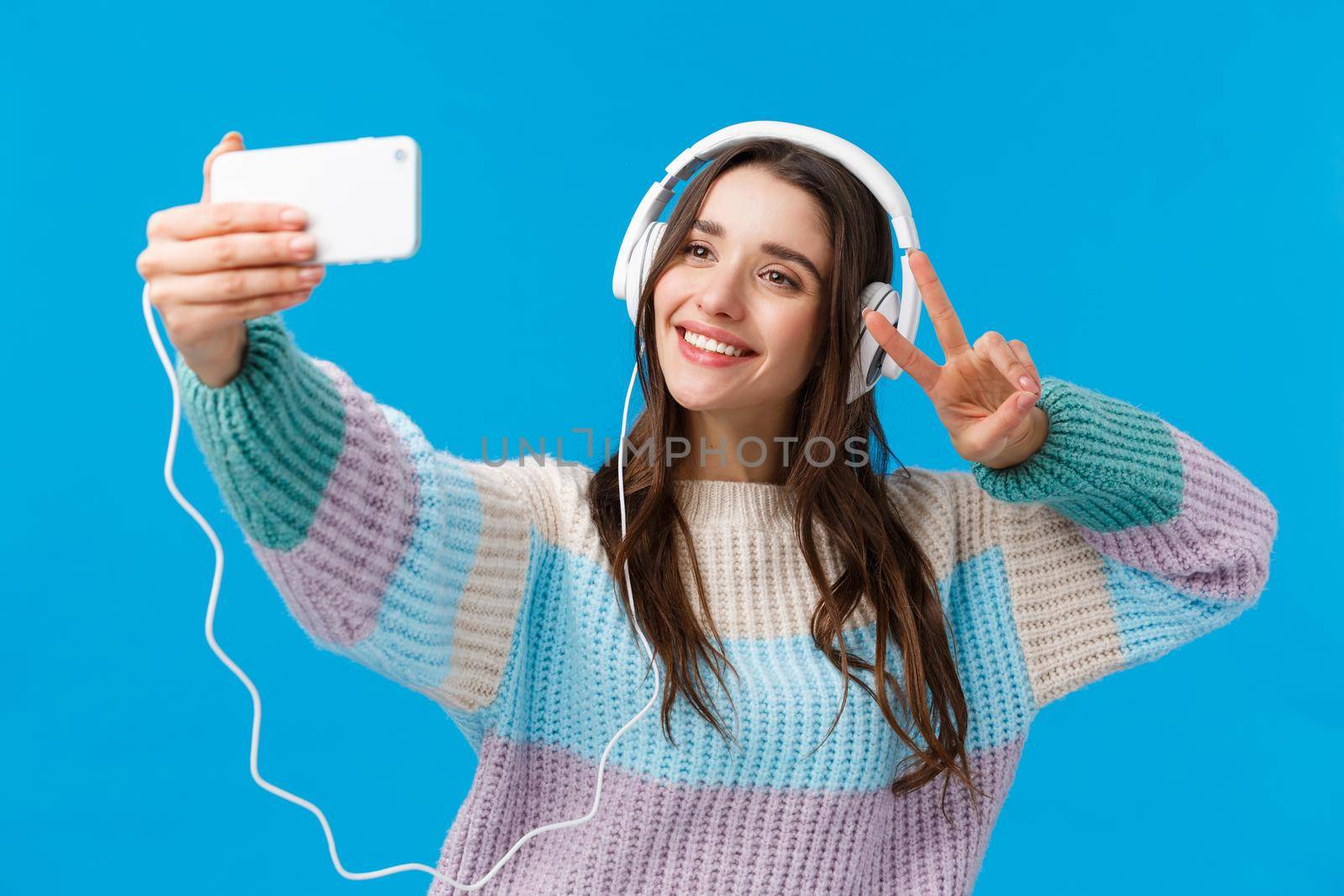 Waist-up portrait cute and tender, lovely charismatic woman taking selfie on smartphone, wearing headphones, listen music, posing with peace gesture as trying-out new app filters for photo.