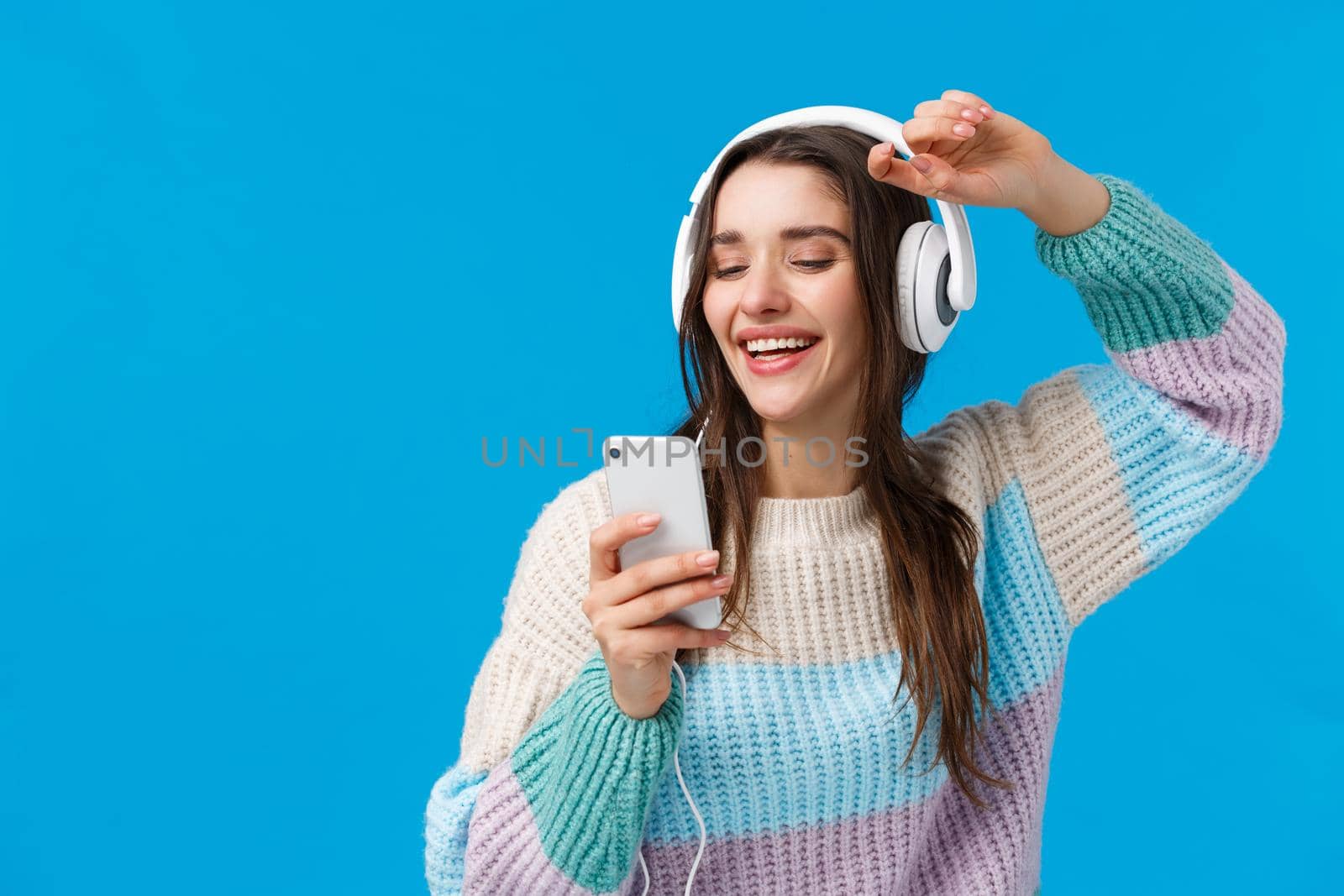 Happy charismatic and carefree smiling european woman enjoying awesome music quality sound in new headphones received as christmas gift, raising hands up holding smartphone, blue background.