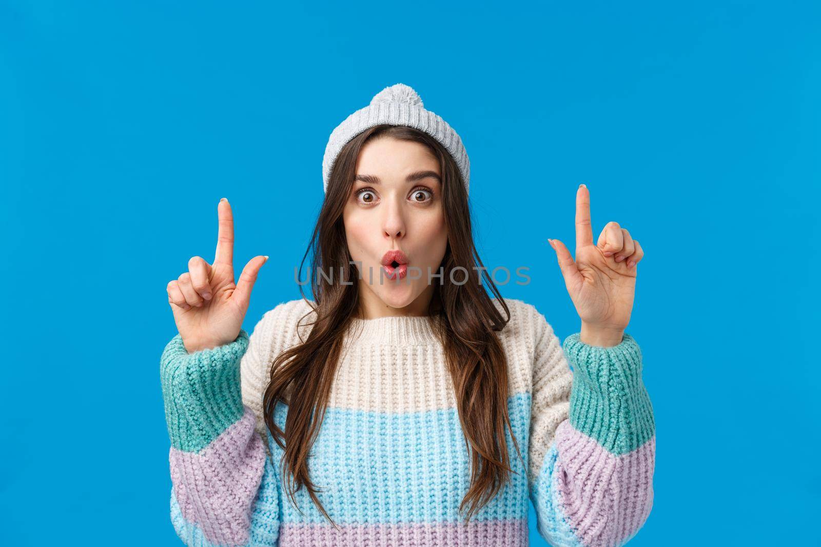 Wow so cool, check this out. Impressed and astonished brunette woman in winter hat, sweater, folding lips amused, looking camera and pointing fingers up, telling about amazing event, blue background by Benzoix