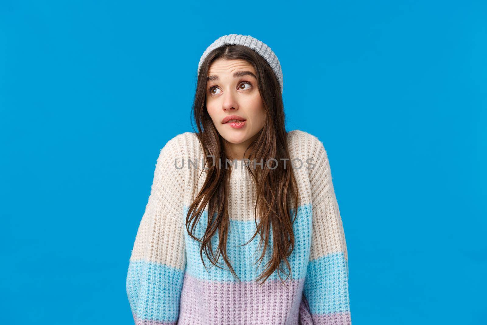 Silly and cute awkward young woman acting uninvolved, biting lip shrugging look away embarrassed, say oops, made mistake avoid eye contact, standing blue background apologizing.