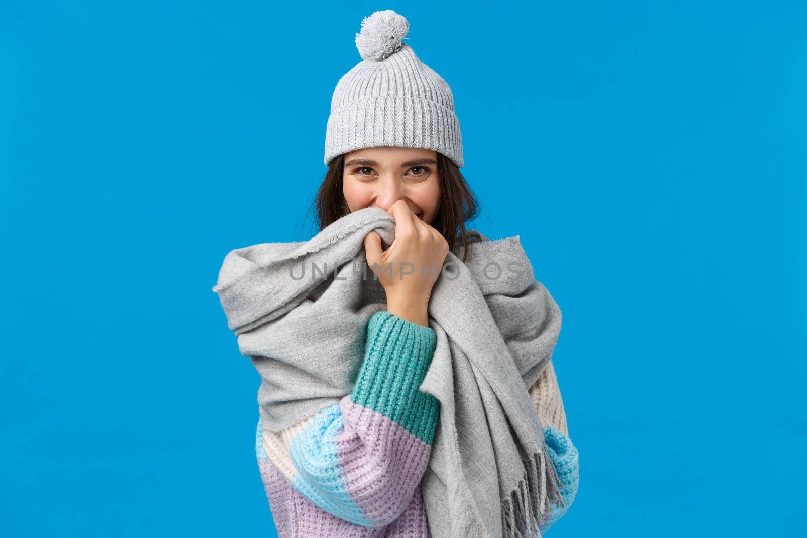 Silly and romantic cute happy woman blushing, hiding smile behind scarf and looking camera joyfully, enjoying romantic st valentines day date, standing blue background cheerful.