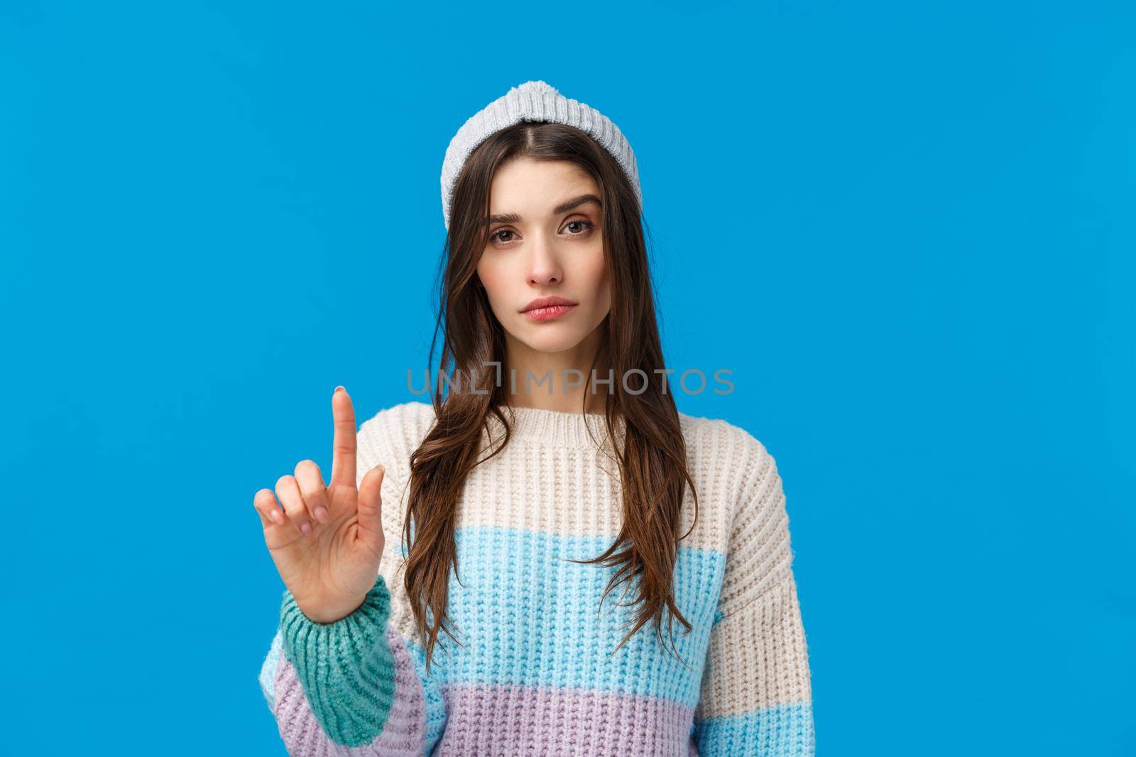 Rule number one. Serious-looking assertive woman in winter sweater, hat, raising one finger in disapproval, restriction or warning, prohibit do something, not so fast gesture, blue background.