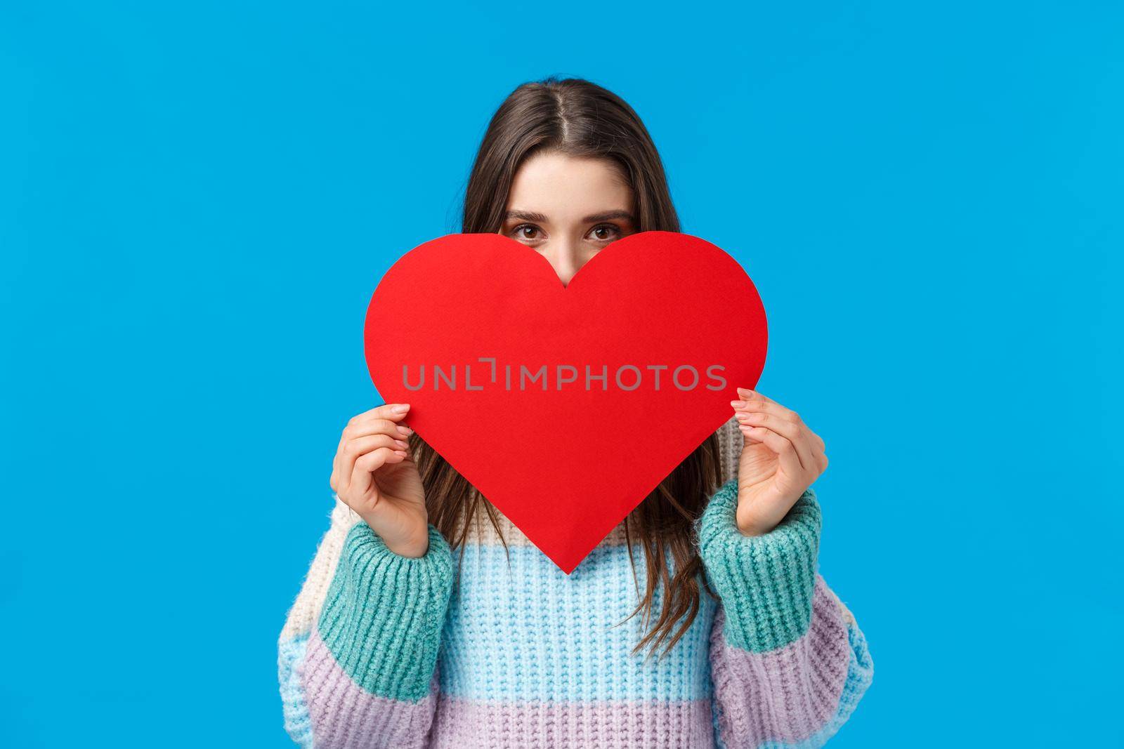 Someone is loved. Blushing cute and silly girlfriend want confess in love, deep feelings, hiding face behind big lovely red heart and looking camera, express affection standing over blue background.
