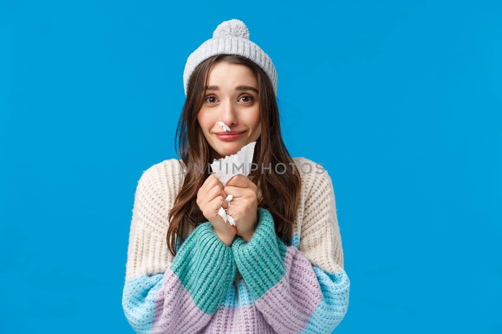 Silly and cute lovely, smiling brunette girl in winter sweater, hat, have runny nose, caught cold on winter holidays, apologizing for sneezing, holding napkin, standing blue background by Benzoix