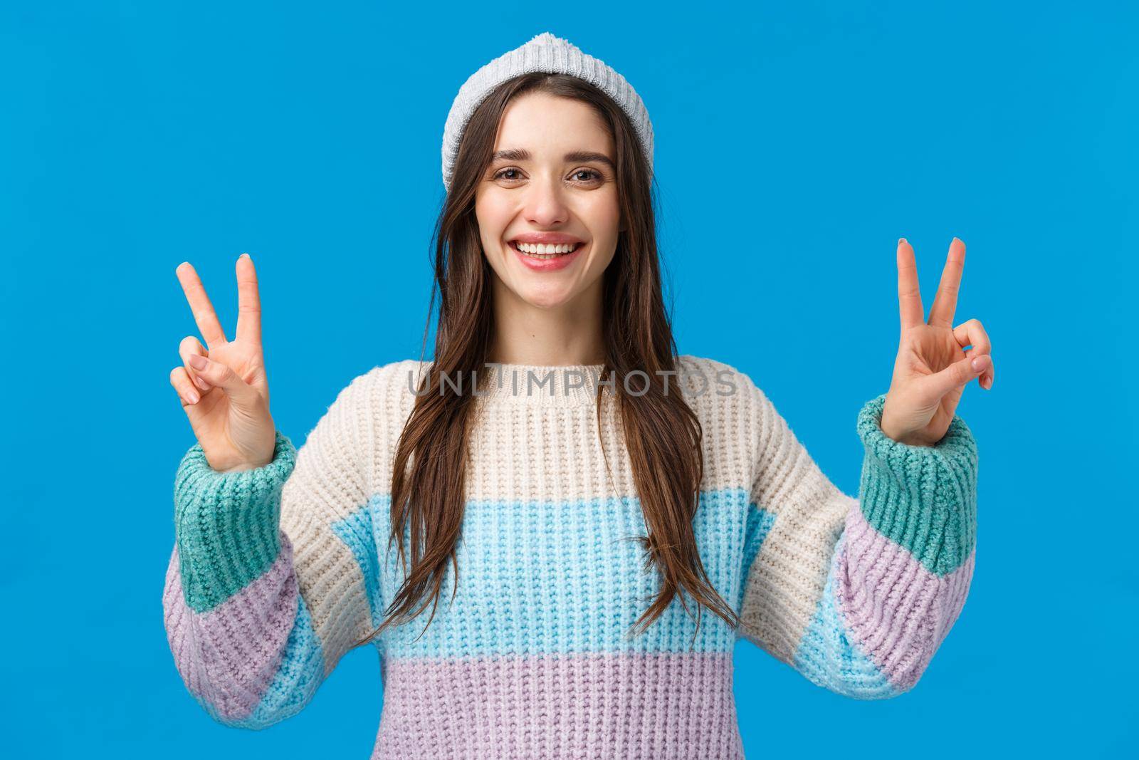 Happy and carefree cute, charismatic brunette female in winter sweater, hat, showing two peace signs and smiling broadly, photographing on winter holidays, mountain ski luxury resort, blue background.