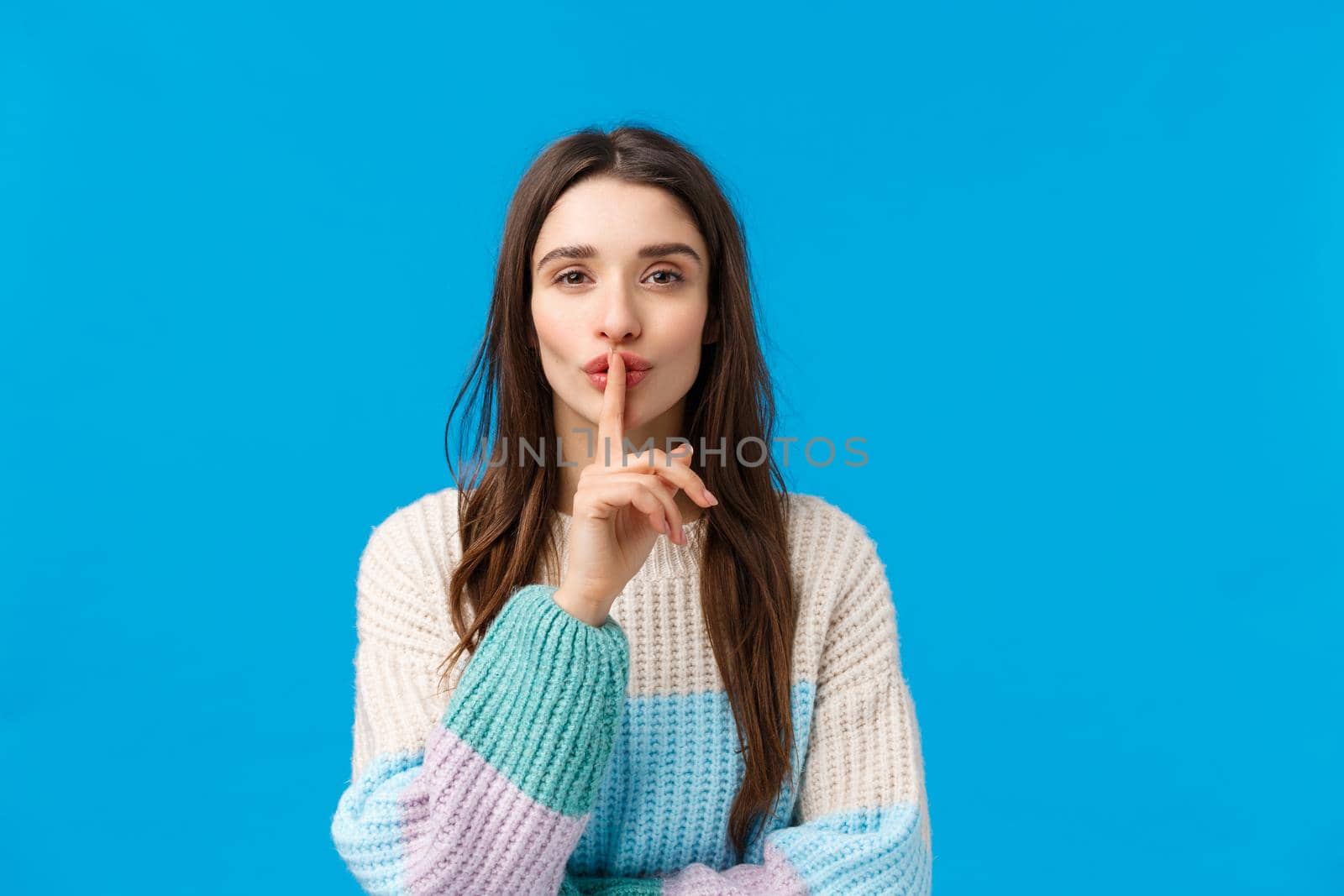 Sensual and cute, attractive young brunette woman in winter sweater, prepare lovely romantic present, hushing smiling and place index finger over mouth, keeping quiet stay silent, blue background by Benzoix