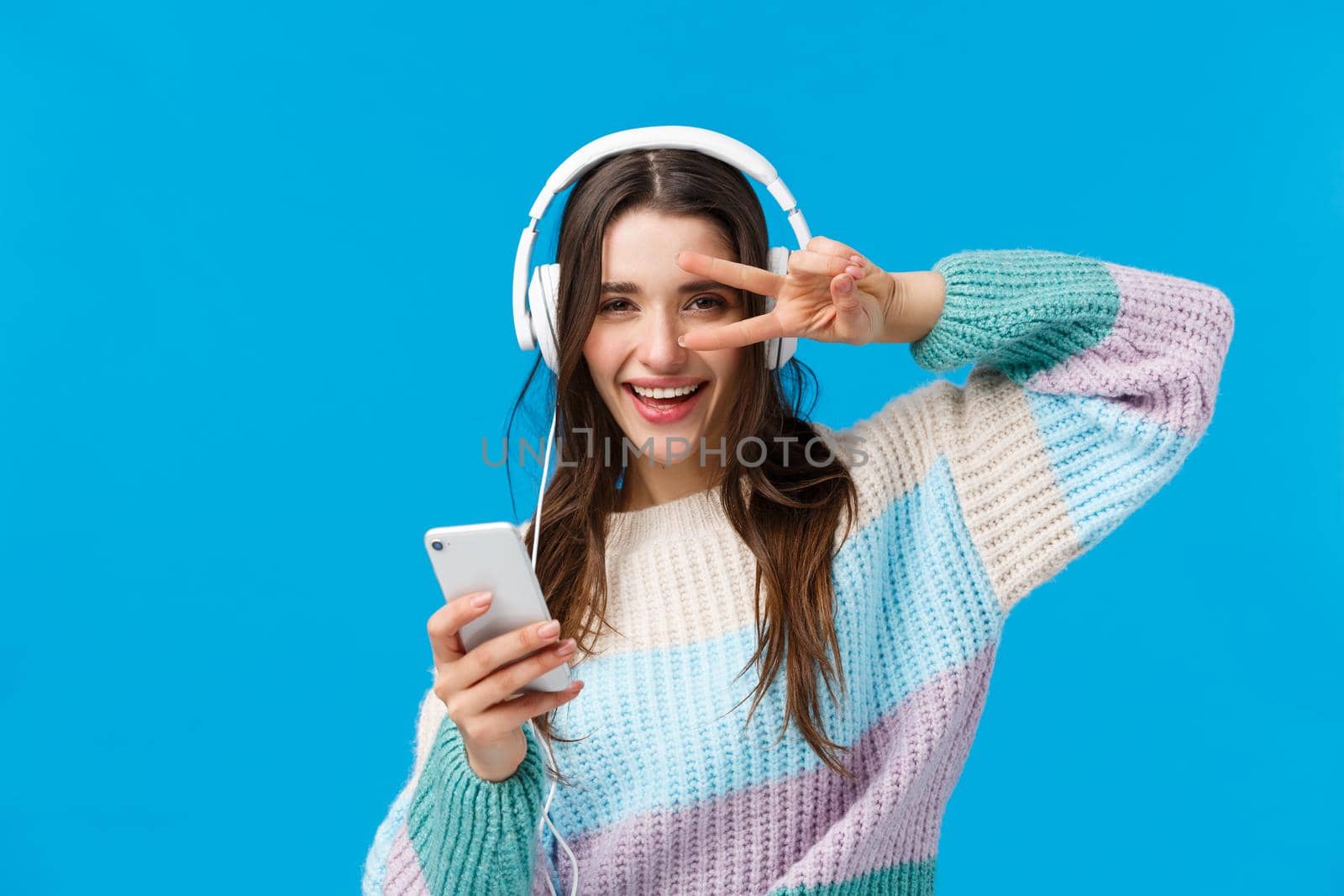 Happy sassy good-looking brunette woman in headphones, winter sweater, enjoying favorite songs in new earphones, show peace, disco gesture holding smartphone, smiling camera pleased.