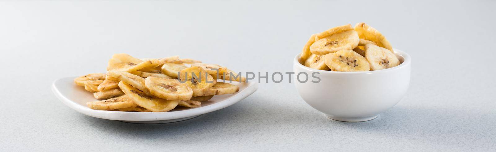 Baked banana chips in a white bowl and saucer on the table. Fast food. Web banner by Aleruana