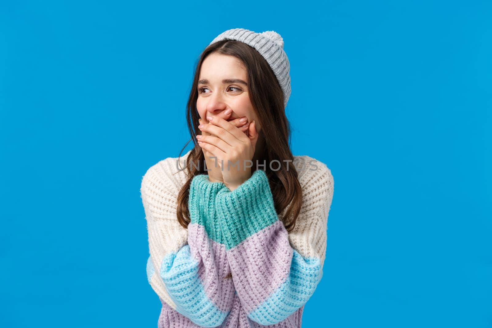 Emotions, fun and happiness concept. Cheerful young caucasian woman in winter sweater, hat, laughing out loud looking aside, laughing close mouth with hands, joking around, standing blue background.