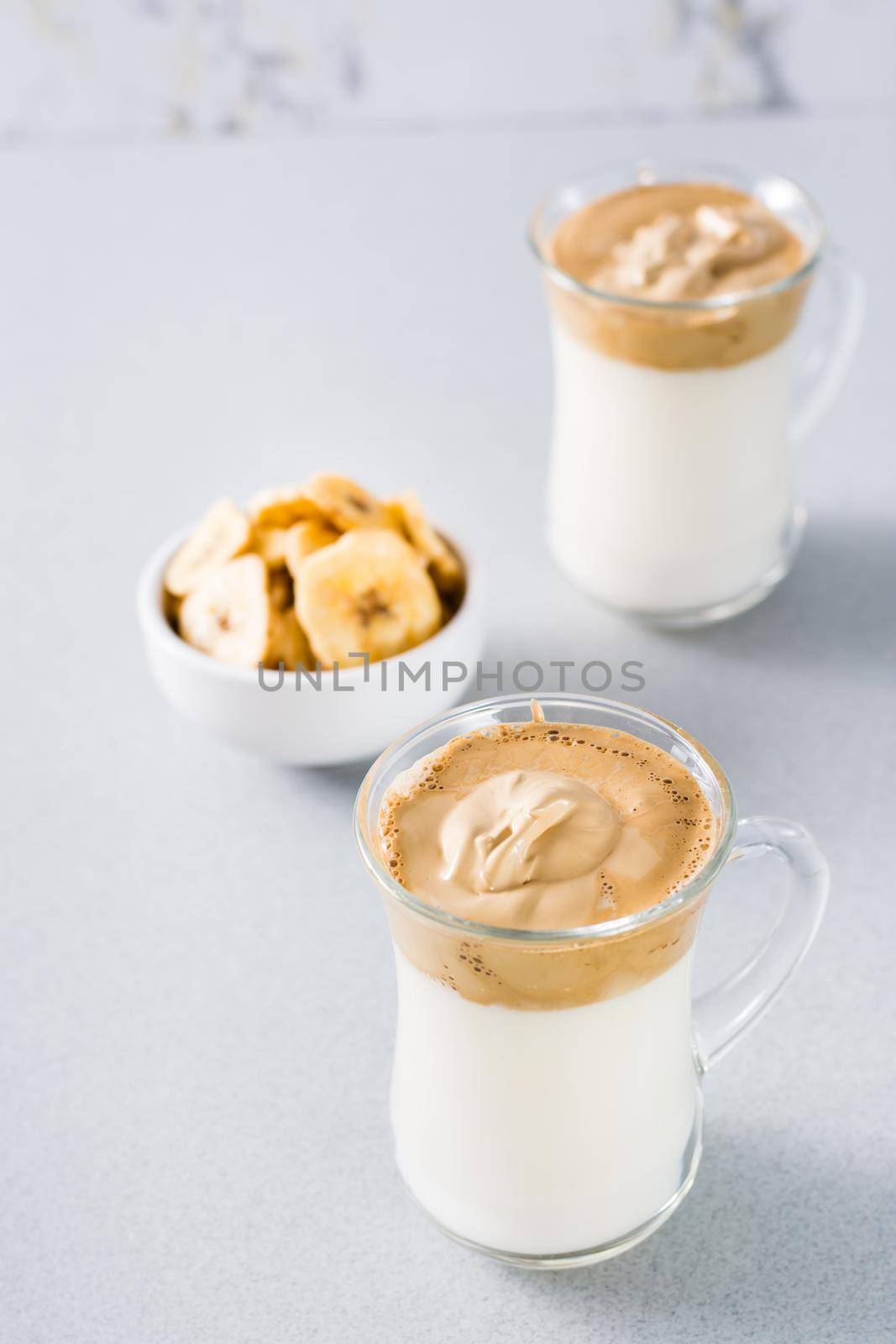 Quarantine trendy cuisine. Two cups with dalgona coffee and banana chips in a bowl on a gray background. Vertical view