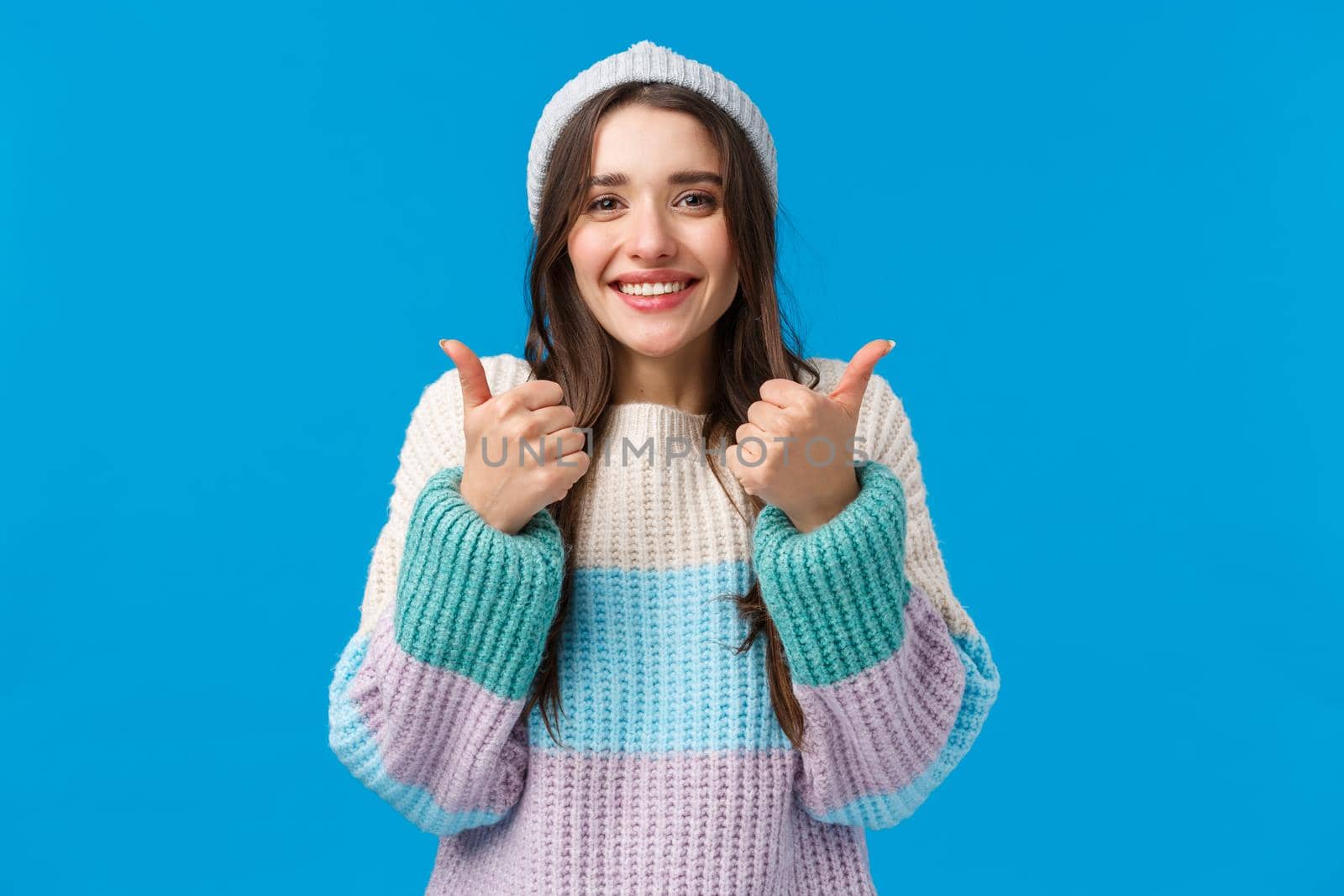 Super good, awesome idea. Cheerful glad cute brunette woman in winter hat, sweater, smiling and showing thumbs-up in approval, encourage friend, say congrats or yes, nod agreement, blue background.