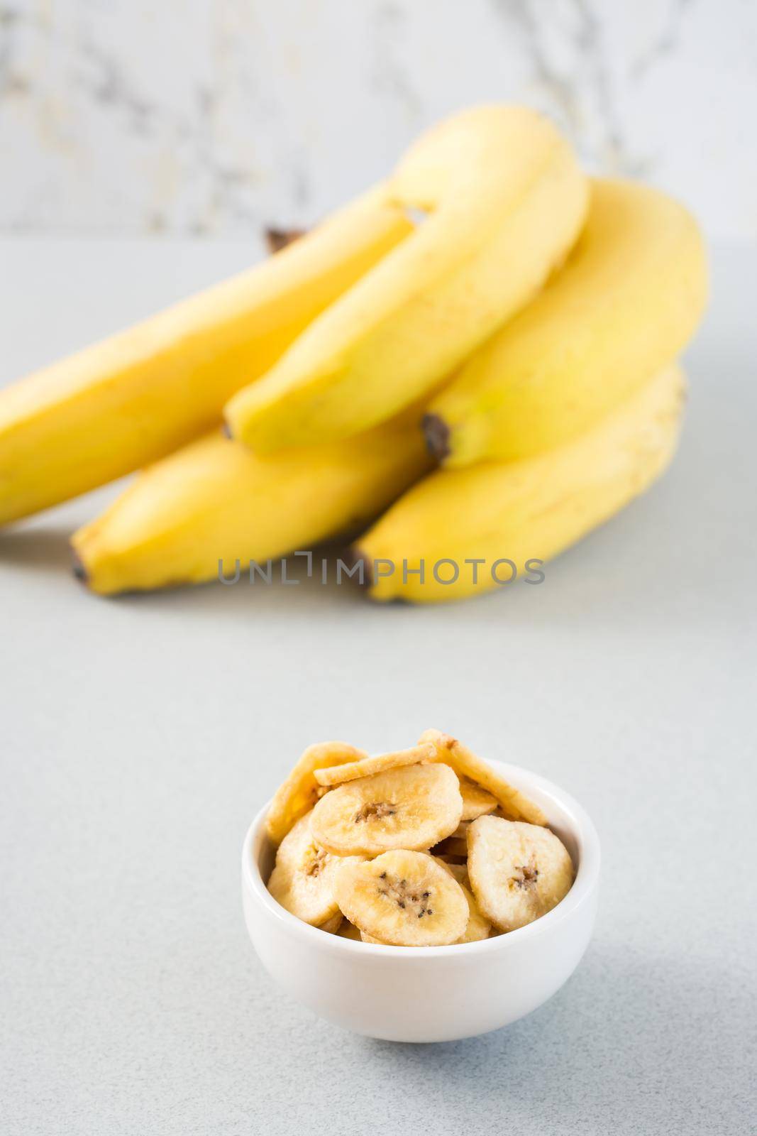 Baked banana chips in a white bowl and a bunch of bananas on the table. Fast food. Close-up. Vertical view by Aleruana