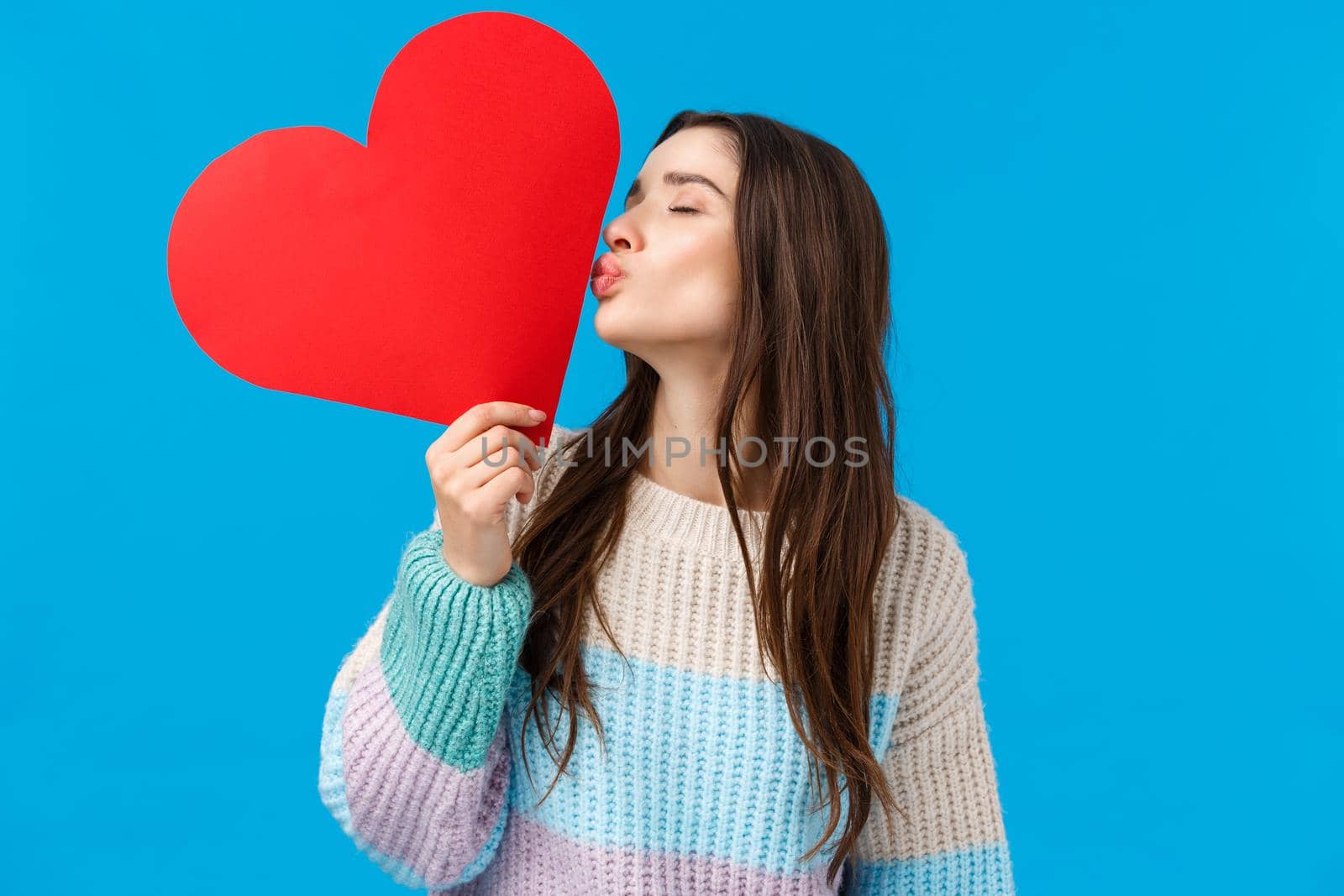 Romance, love and tenderness concept. Feminine cheerful and cute brunette woman showing her feelings, kissing big red heart card with closed eyes, express sympathy and care, blue background.