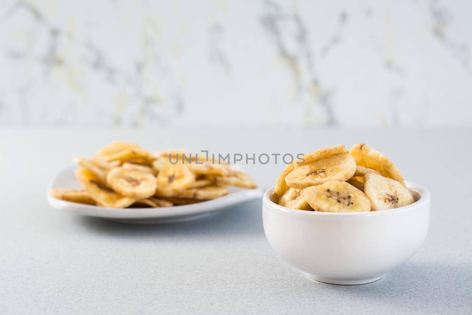 Baked banana chips in a white bowl and saucer on the table. Fast food. Copy space by Aleruana