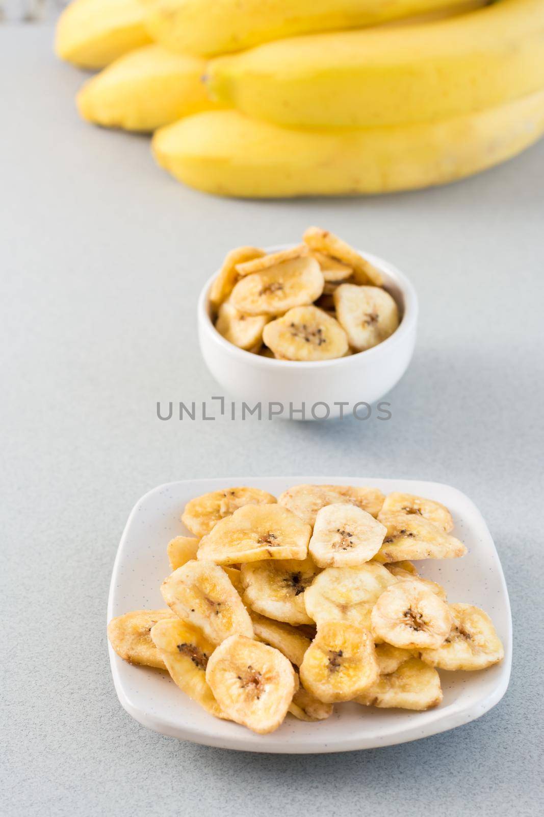 Baked banana chips in a white bowl and saucer and a bunch of bananas on the table. Fast food. Vertical view by Aleruana
