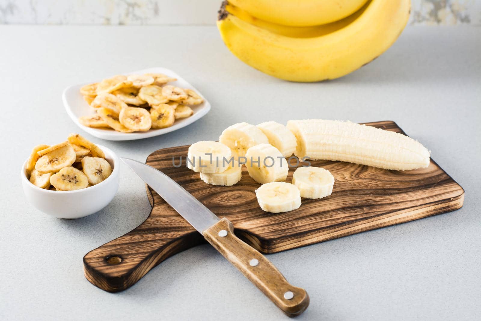 Banana slices on a cutting board and a knife and banana chips in a bowl on the table. Fast food. by Aleruana