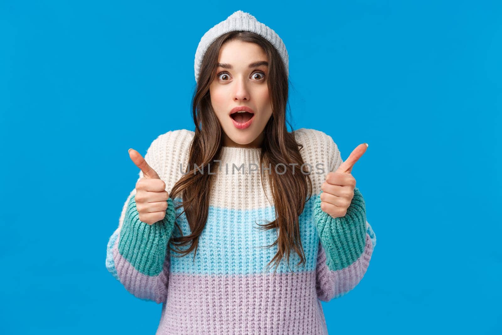 Surprised and happy amazed young brunette woman in winter hat, sweater, stare camera impressed, gasping hold breath speechless staring awesome thing and show thumbs-up in like, yes gesture by Benzoix
