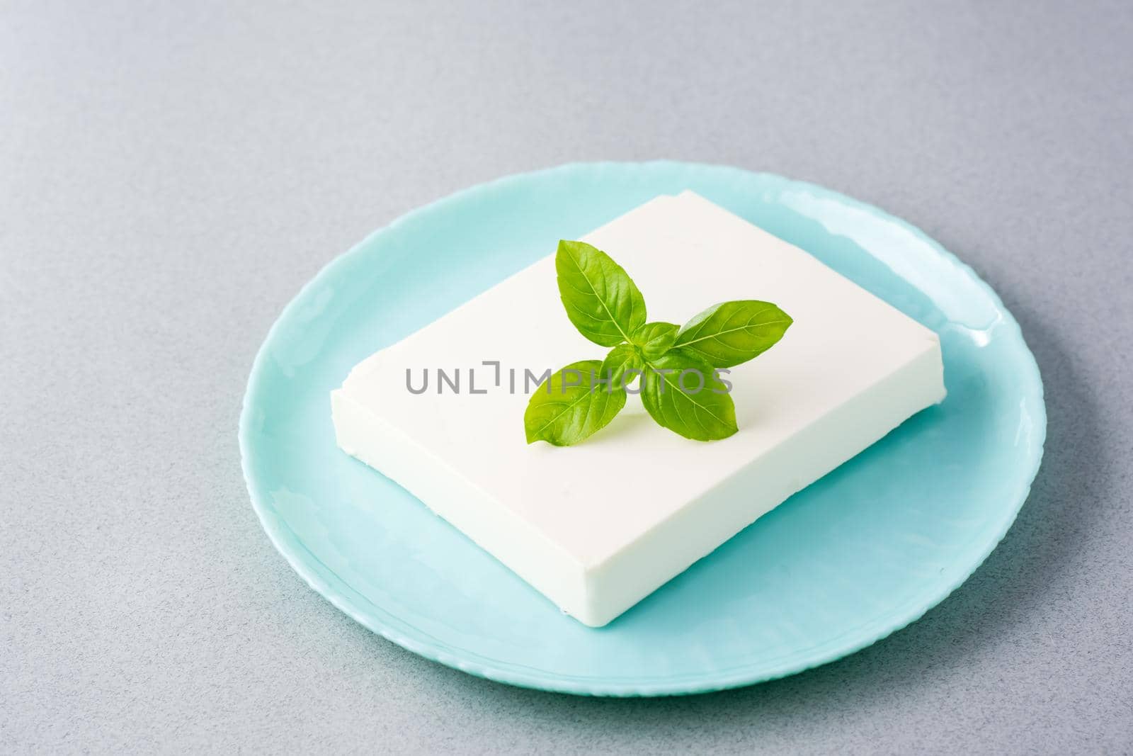 A piece of fresh feta cheese and basil leaves on a plate on the table. Ingredient for Greek salad