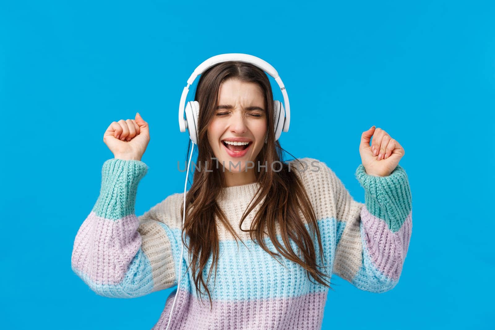 Rock n roll baby. Carefree emotive charismatic brunette woman enjoying listening music in new gift headphones, dancing raising hands up, close eyes and singing along favorite song, blue background.