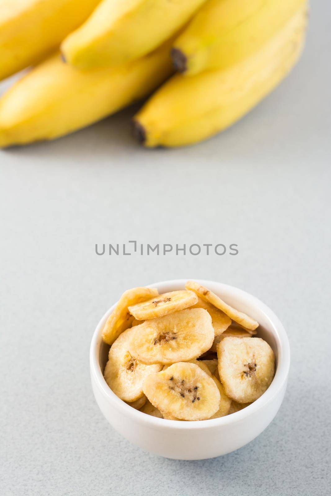 Baked banana chips in a white bowl and a bunch of bananas on the table. Fast food. Copy space. Vertical view by Aleruana