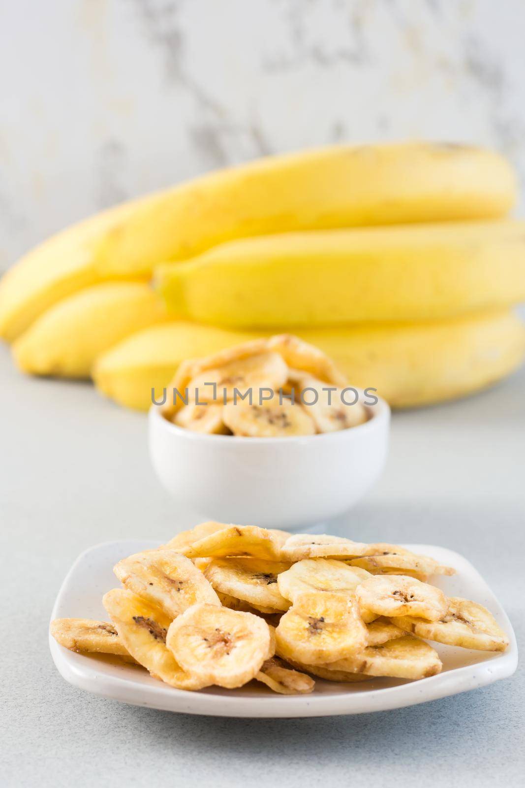 Baked banana chips in a white bowl and saucer and a bunch of bananas on the table. Fast food. Vertical view. Close-up
