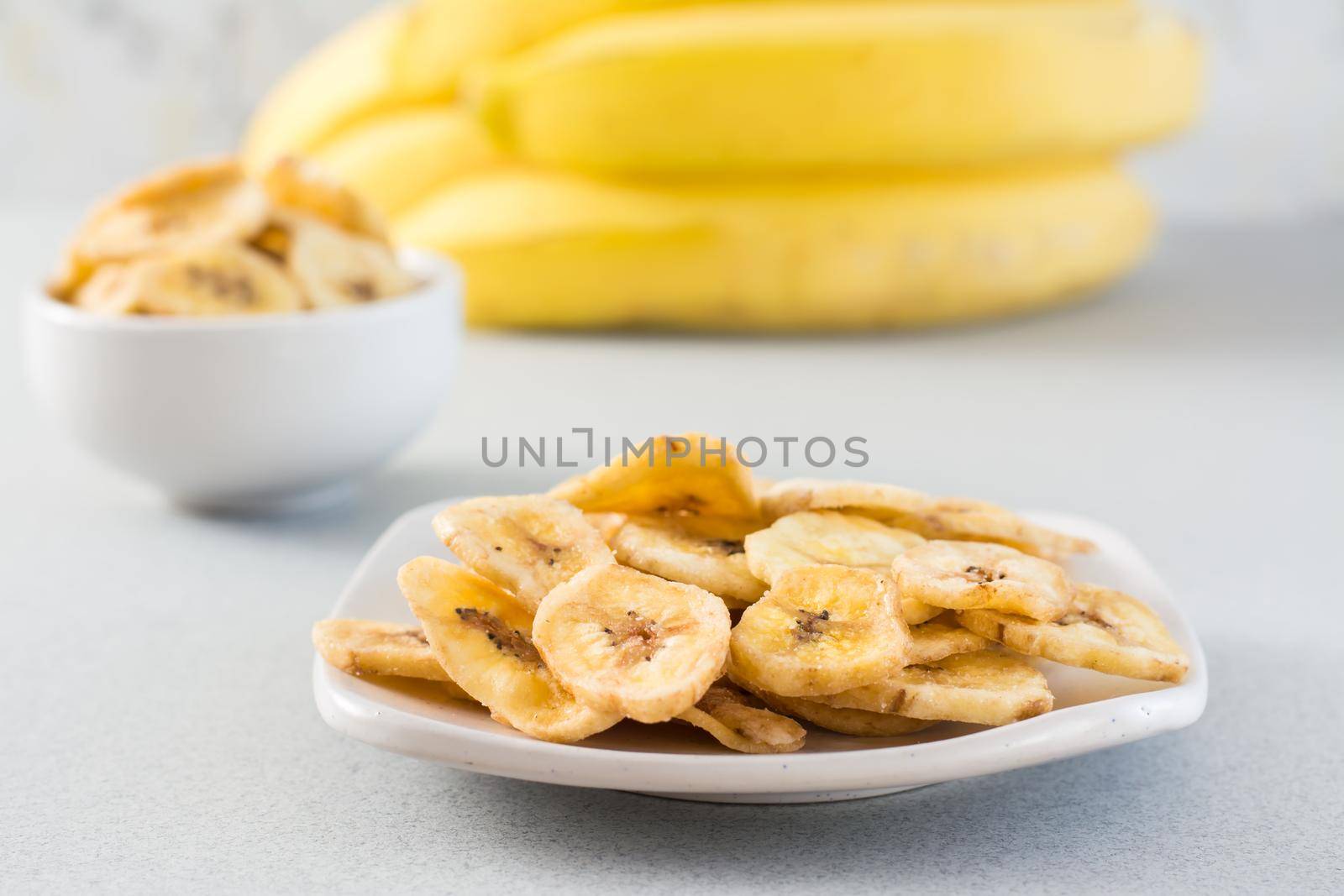 Baked banana chips in a white bowl and saucer and a bunch of bananas on the table. Fast food.
