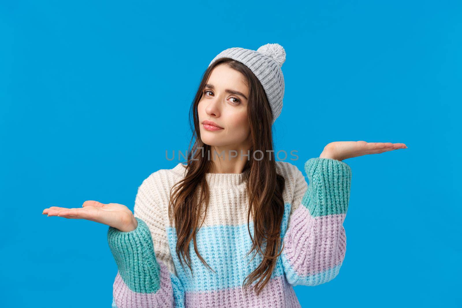 Girl troubled make choice, finally decided what buy, holding hands raised like weighing products, looking camera thoughtful, indecisive, asking help with picking winter holidays gift, blue background.