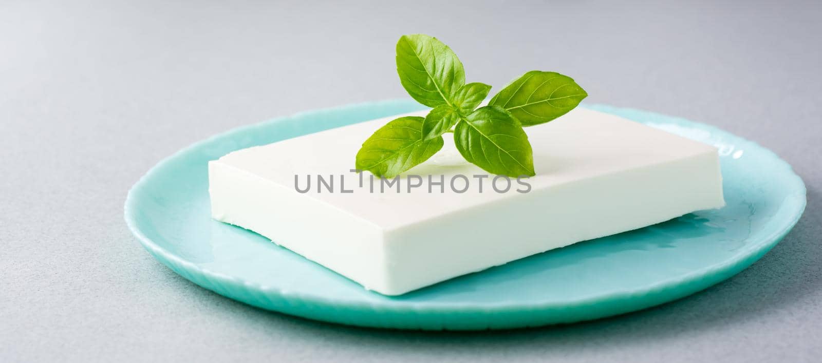 A piece of fresh feta cheese and basil leaves on a plate on the table. Ingredient for Greek salad. Web banner by Aleruana