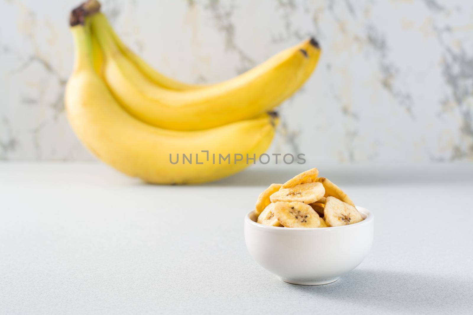 Baked banana chips in a white bowl and a bunch of bananas on the table. Fast food. Copy space