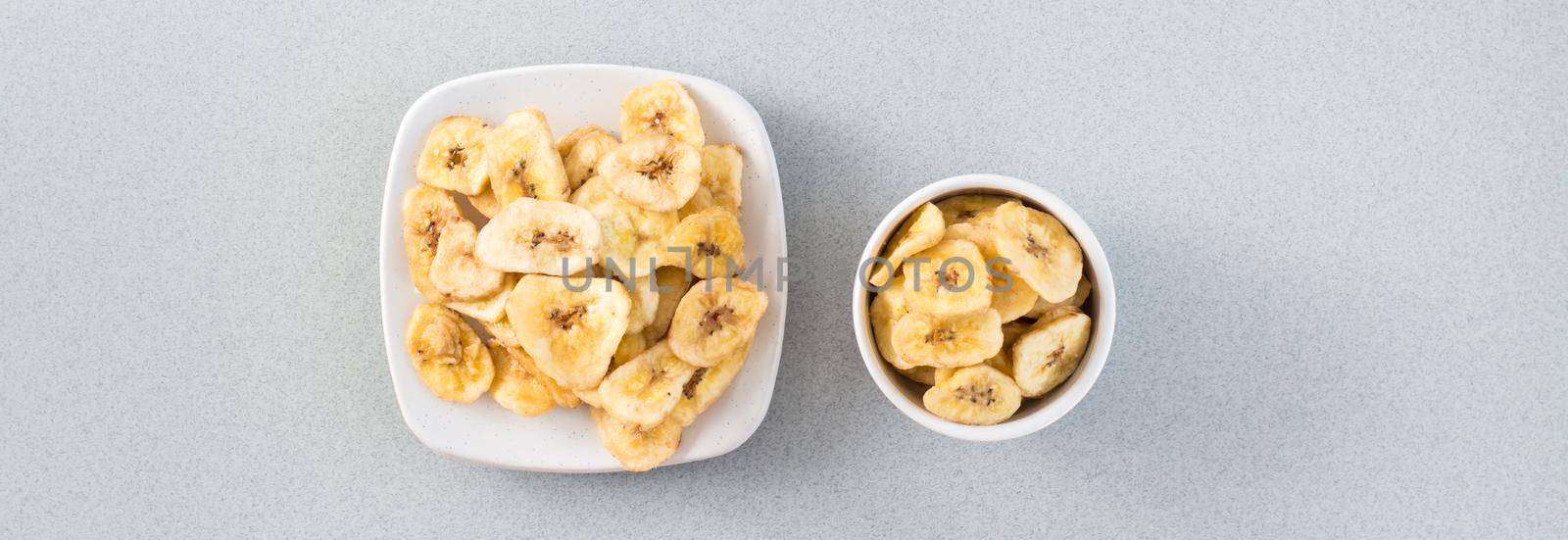 Baked banana chips in a white bowl and saucer on the table. Fast food. Web banner. Top view by Aleruana