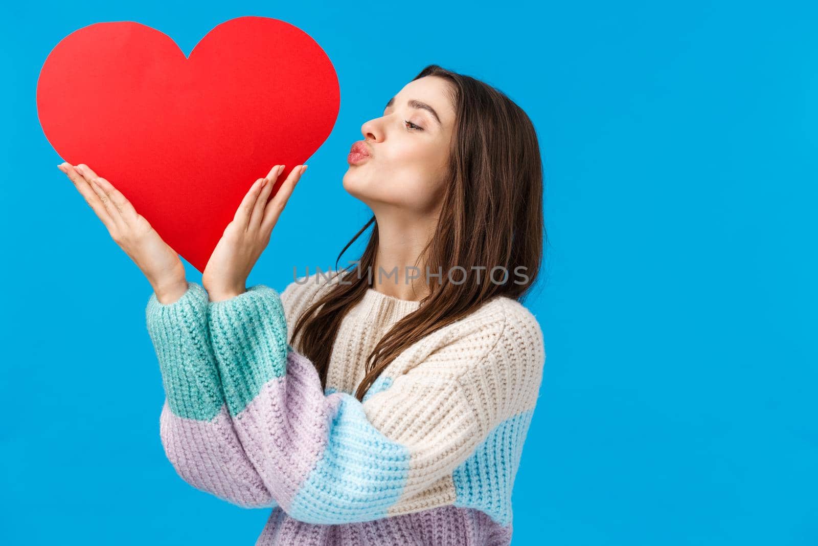 Dreamy young woman cherish her relationship, prepare valentines day gift, kissing big cute red heart sign over left side copy space, standing blue background delighted and upbeat.