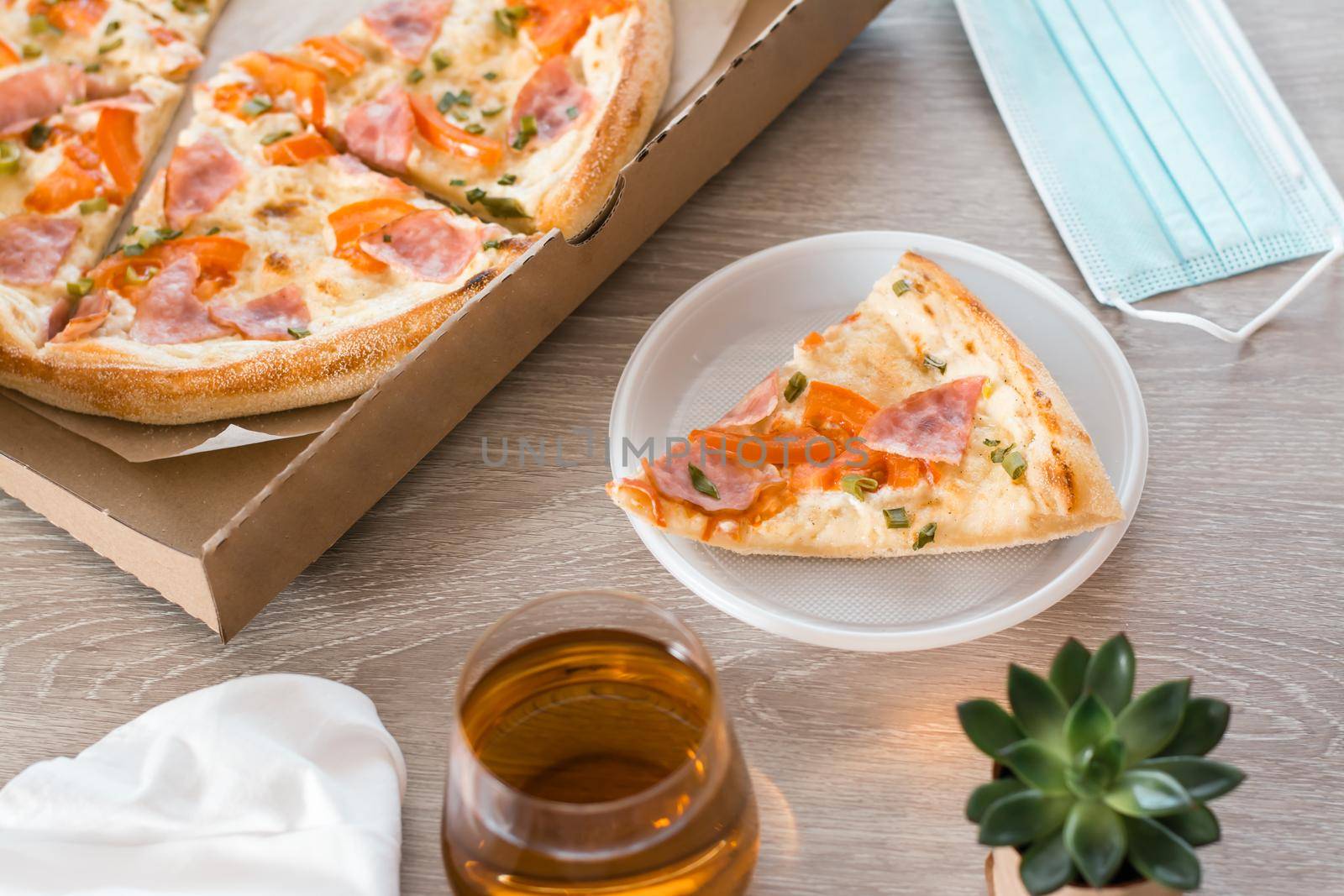 Takeout food. A slice of pizza in a disposable plastic plate, protective mask and a box of pizza on the table in the kitchen.