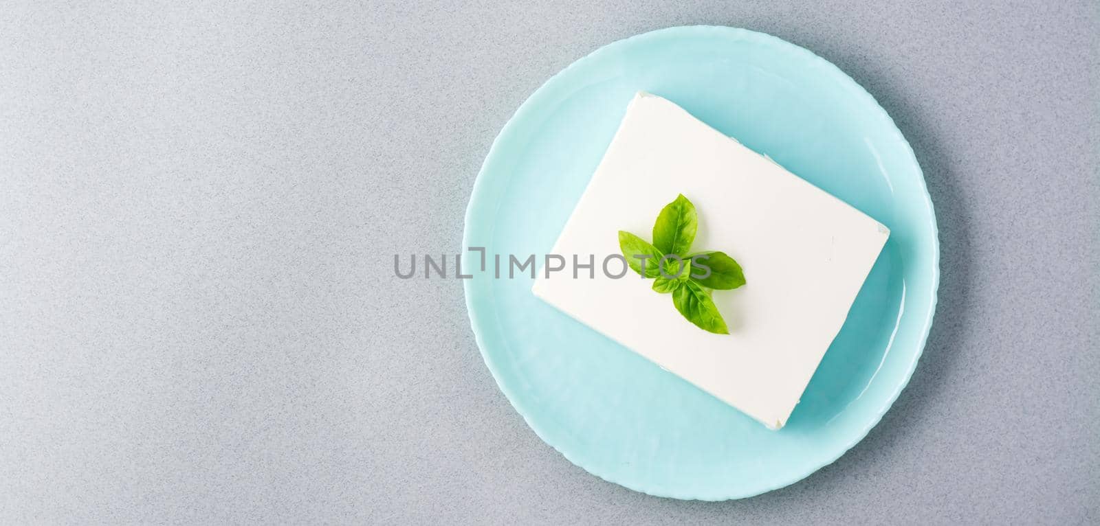 A piece of fresh feta cheese and basil leaves on a plate on the table. Ingredient for Greek salad. Top view. Web banner. Copy space by Aleruana