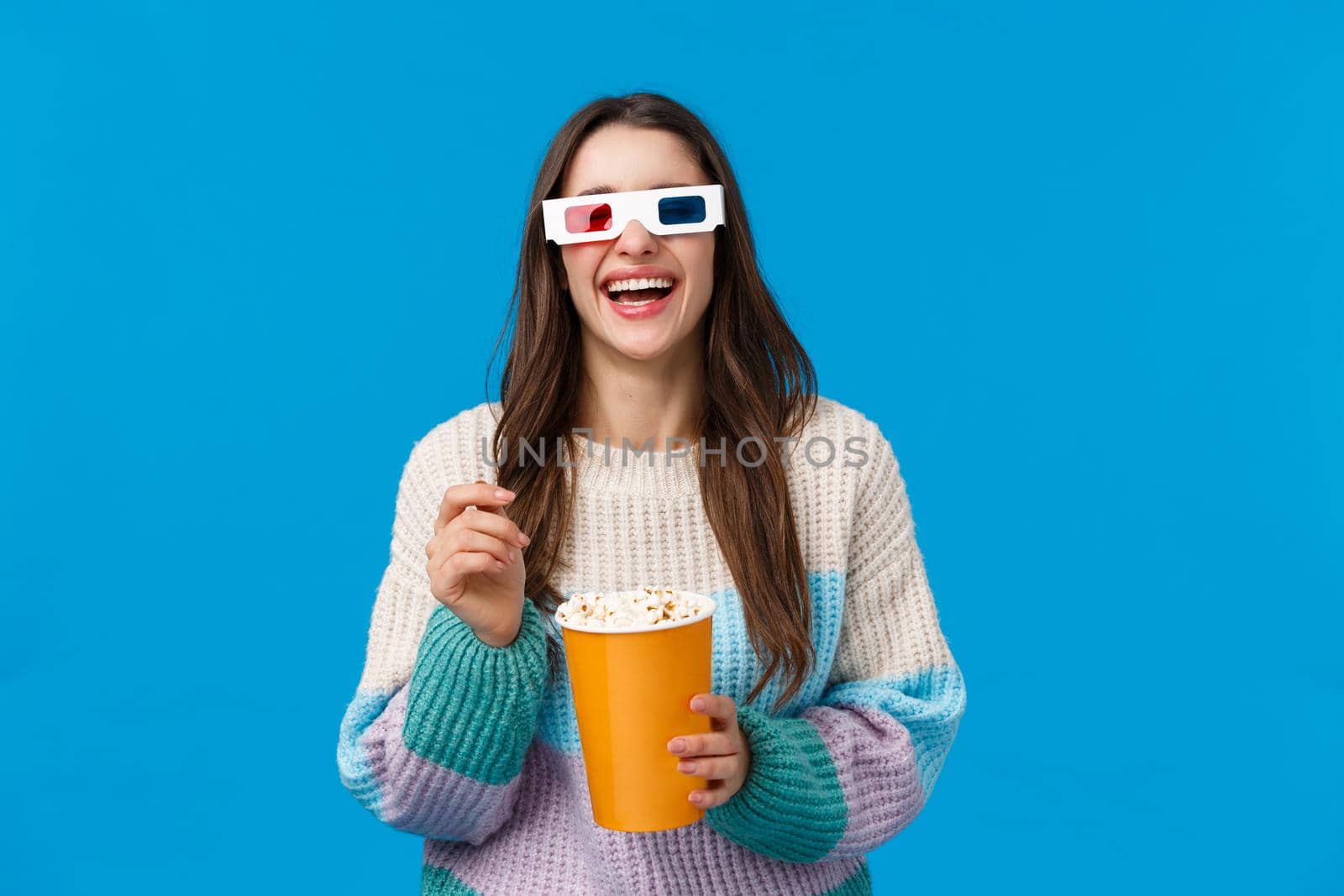 Girl watching comedy in cinema. Carefree happy young brunette woman in winter sweater, enjoying funny movie, laughing out loud eating popcorn, wearing 3d glasses, standing blue background.