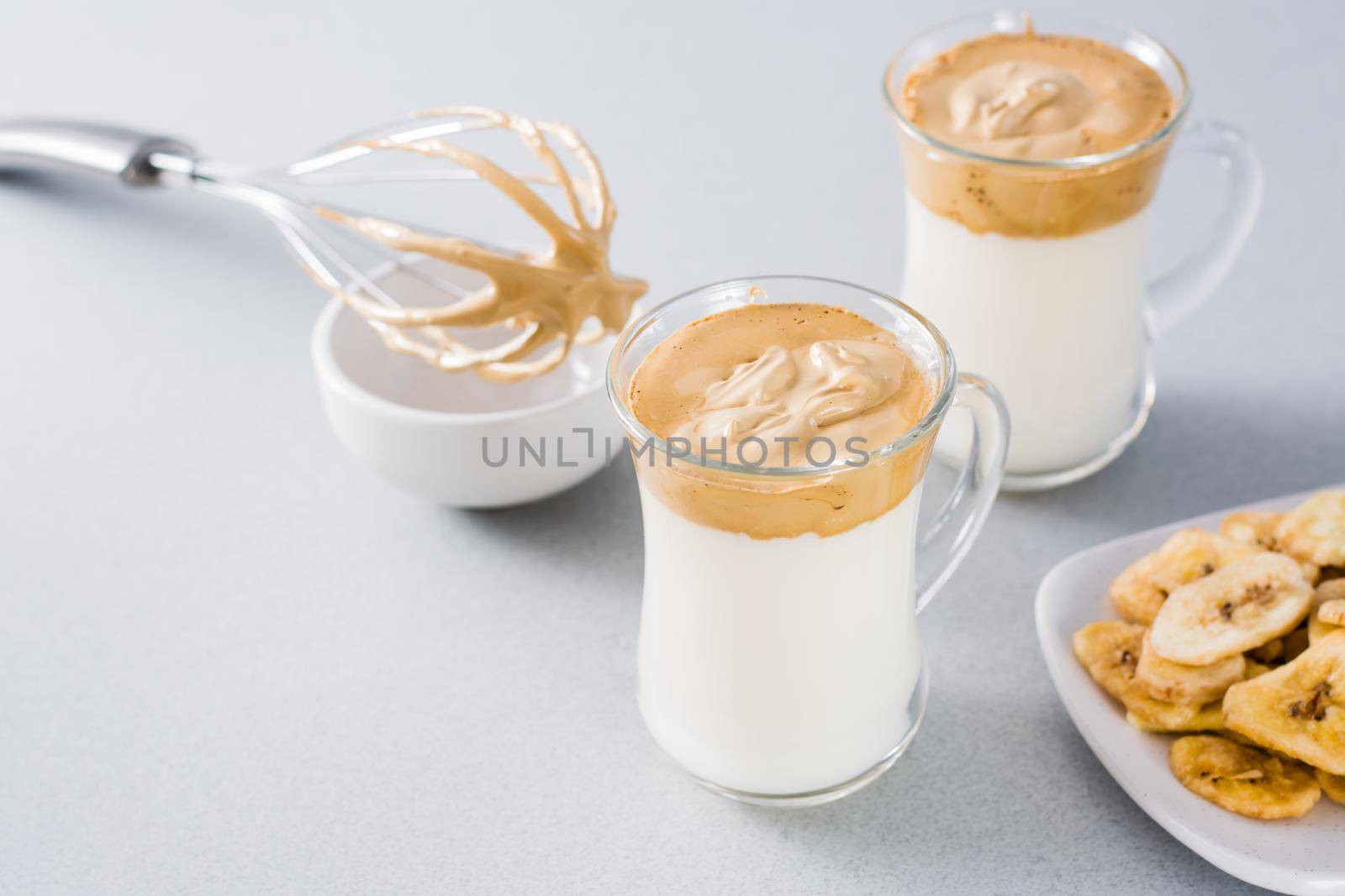 Quarantine trendy cuisine. Two cups with dalgona coffee and banana chips and a whisk on a gray background. by Aleruana