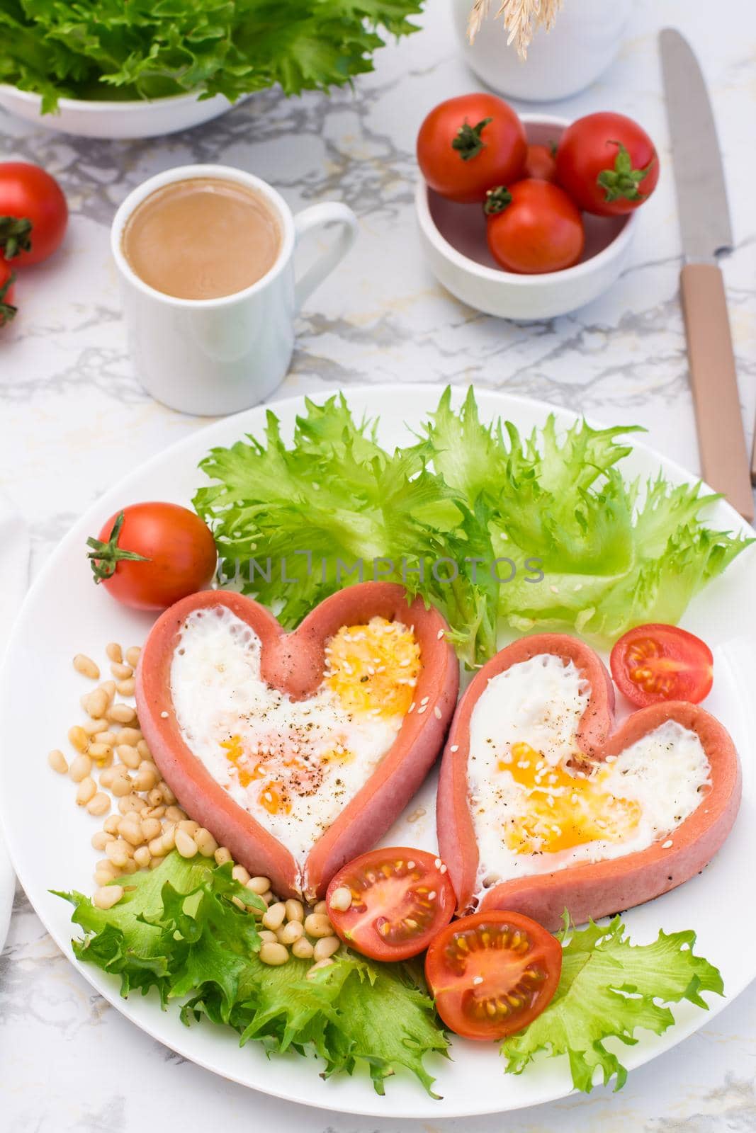 Love breakfast. Fried eggs in heart shaped sausages, lettuce and cherry tomatoes on a plate and a cup of coffee. Vertical view by Aleruana
