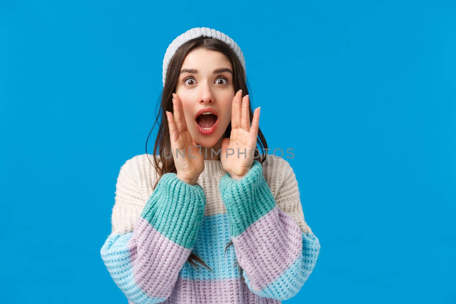 Waist-up portrait silly, cute feminine young woman in winter hat, sweater, calling for someone, shouting with hands near opened mouth, looking camera searching, standing blue background.