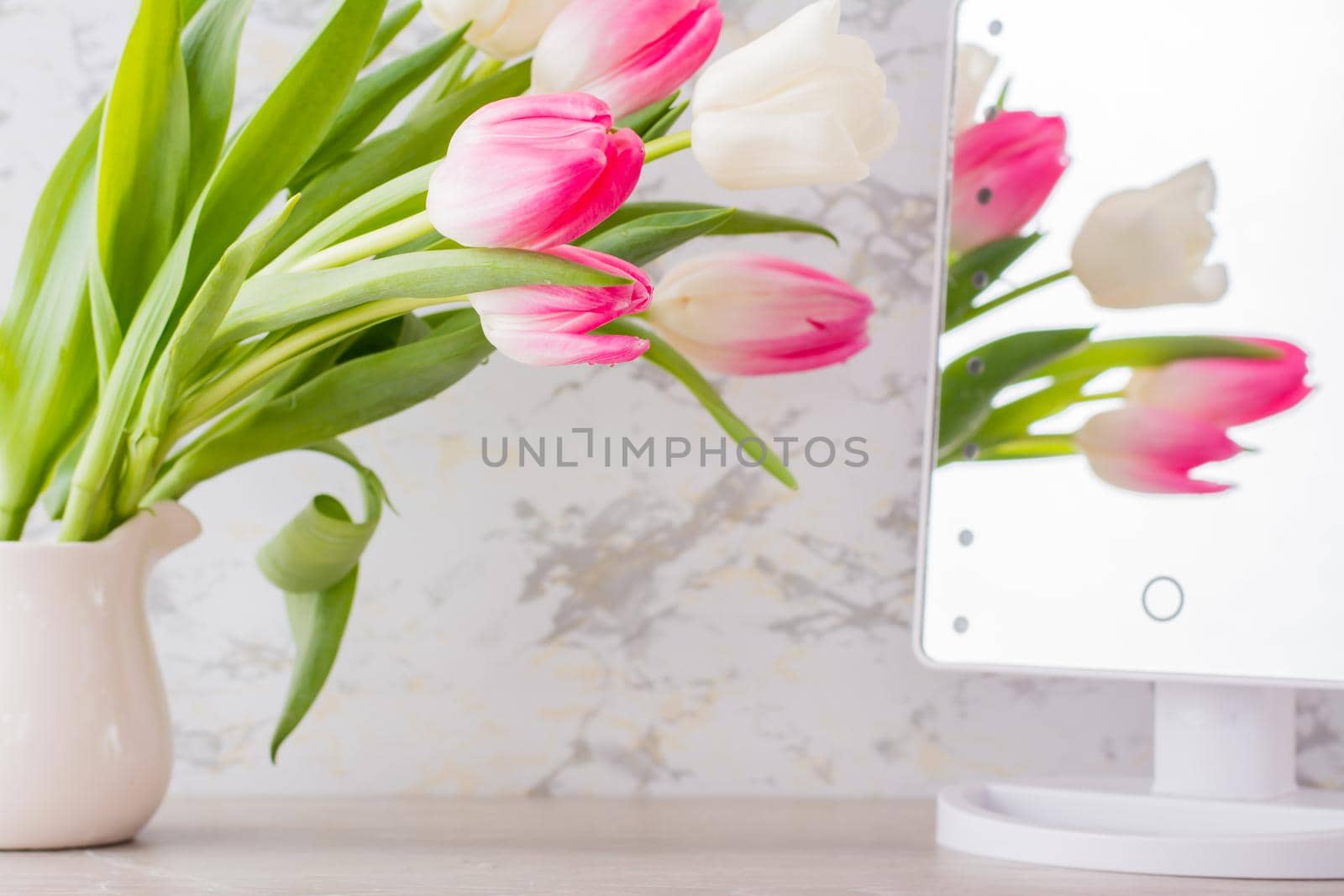 A bouquet of white and pink tulips with green leaves stands in a jug on the table and is reflected in the mirror. Selective focus. by Aleruana