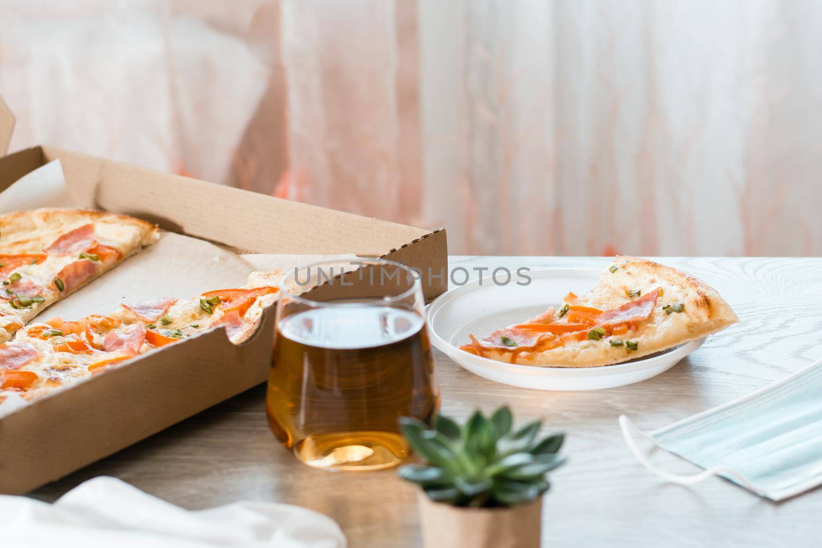 Takeout food. A slice of pizza in a disposable plastic plate and a box of pizza on the table in the kitchen.