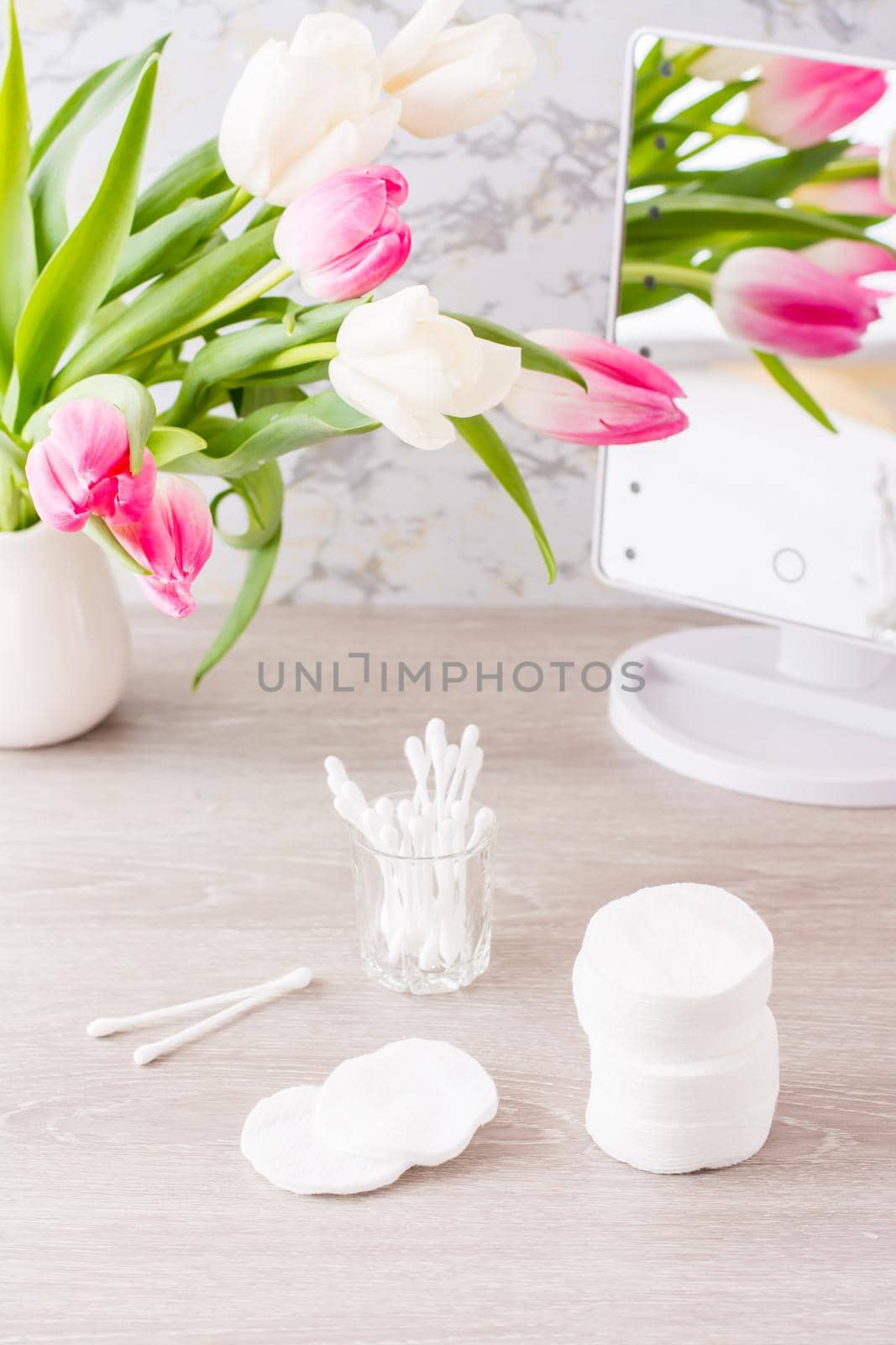 Personal hygiene, cleanliness and skin care. Cotton pads and swabs in a glass on a table in front of a mirror and a bouquet of tulips in a vase. Vertical view by Aleruana
