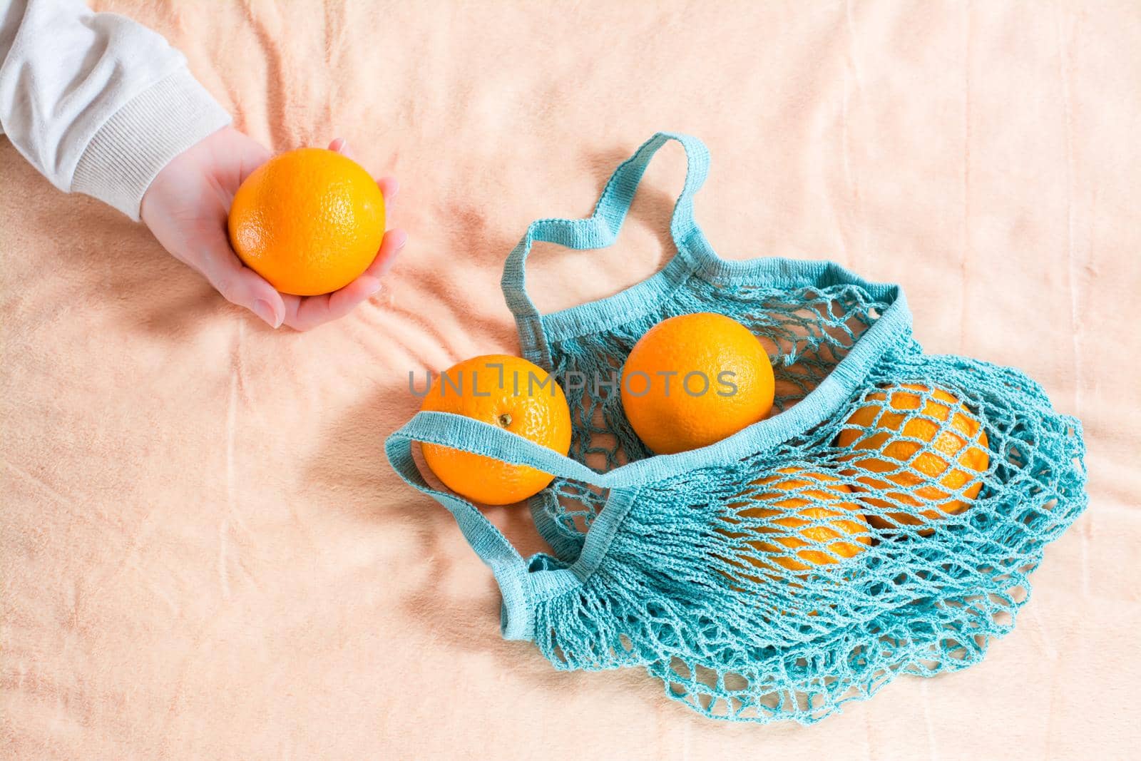 A female hand pulls out fresh oranges from a mesh bag on a fabric background. Zero waste by Aleruana
