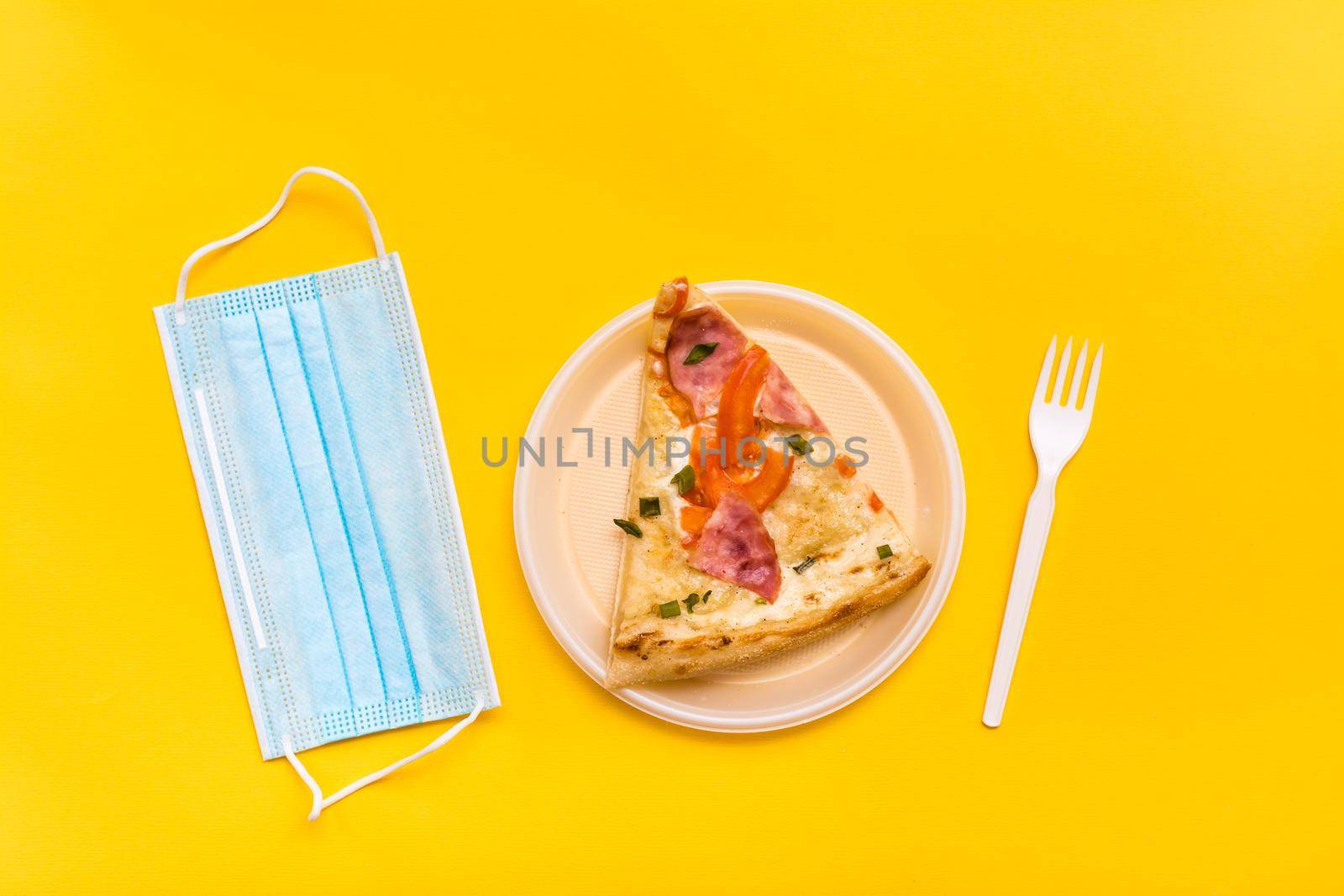 Takeaway and delivery. A slice of pizza in a disposable plastic plate, a protective mask and a fork on a yellow background